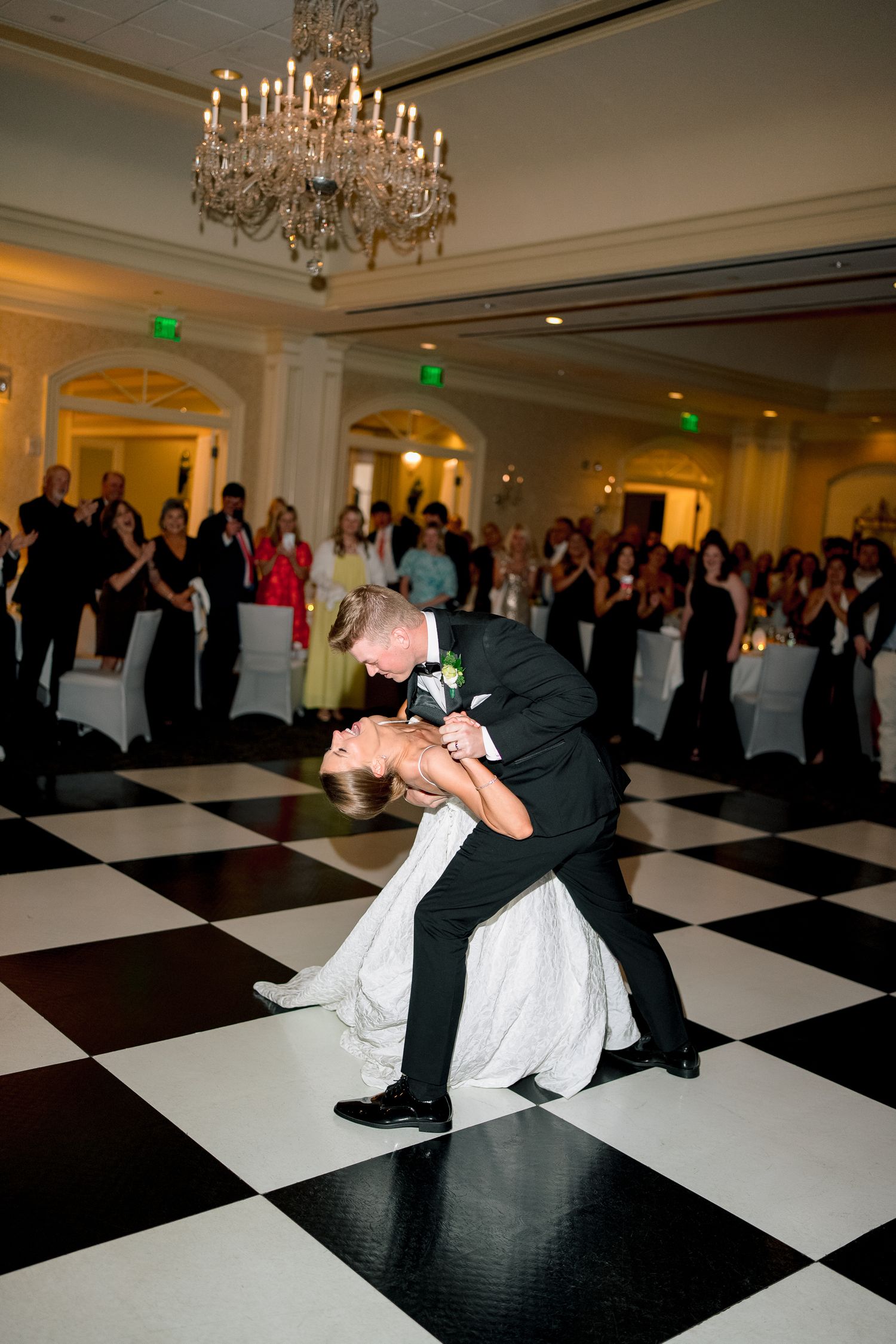 Bride and groom share first dance dip on black and white checkered floor in elegant ballroom with crystal chandelier.