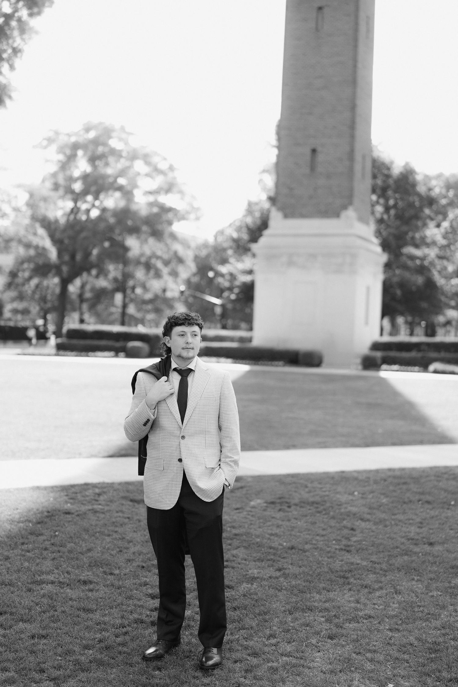 Young man in suit and tie standing on campus lawn with monument tower in black and white photo.