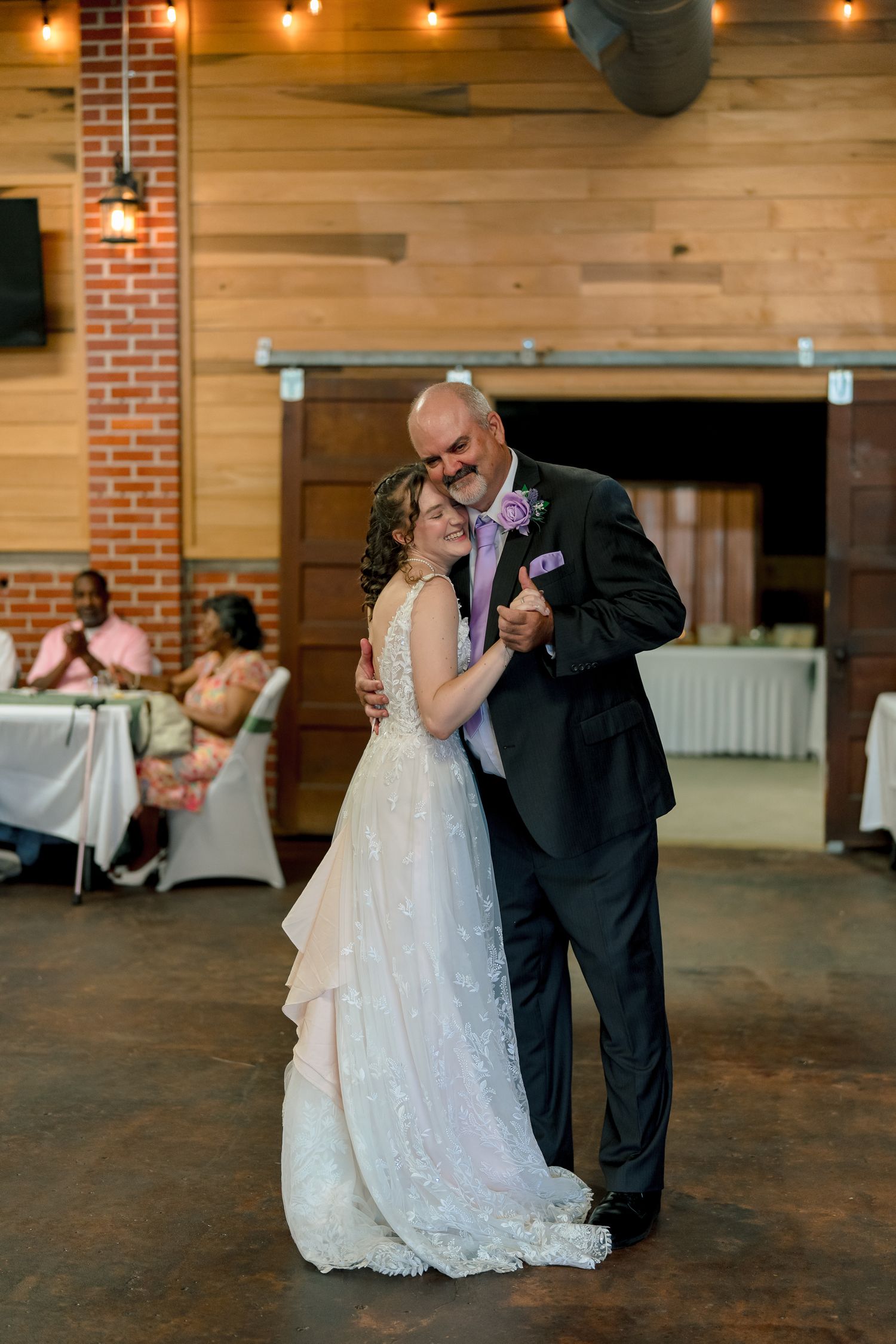 Bride and groom sharing romantic first dance in rustic wedding reception venue with string lights and brick walls.
