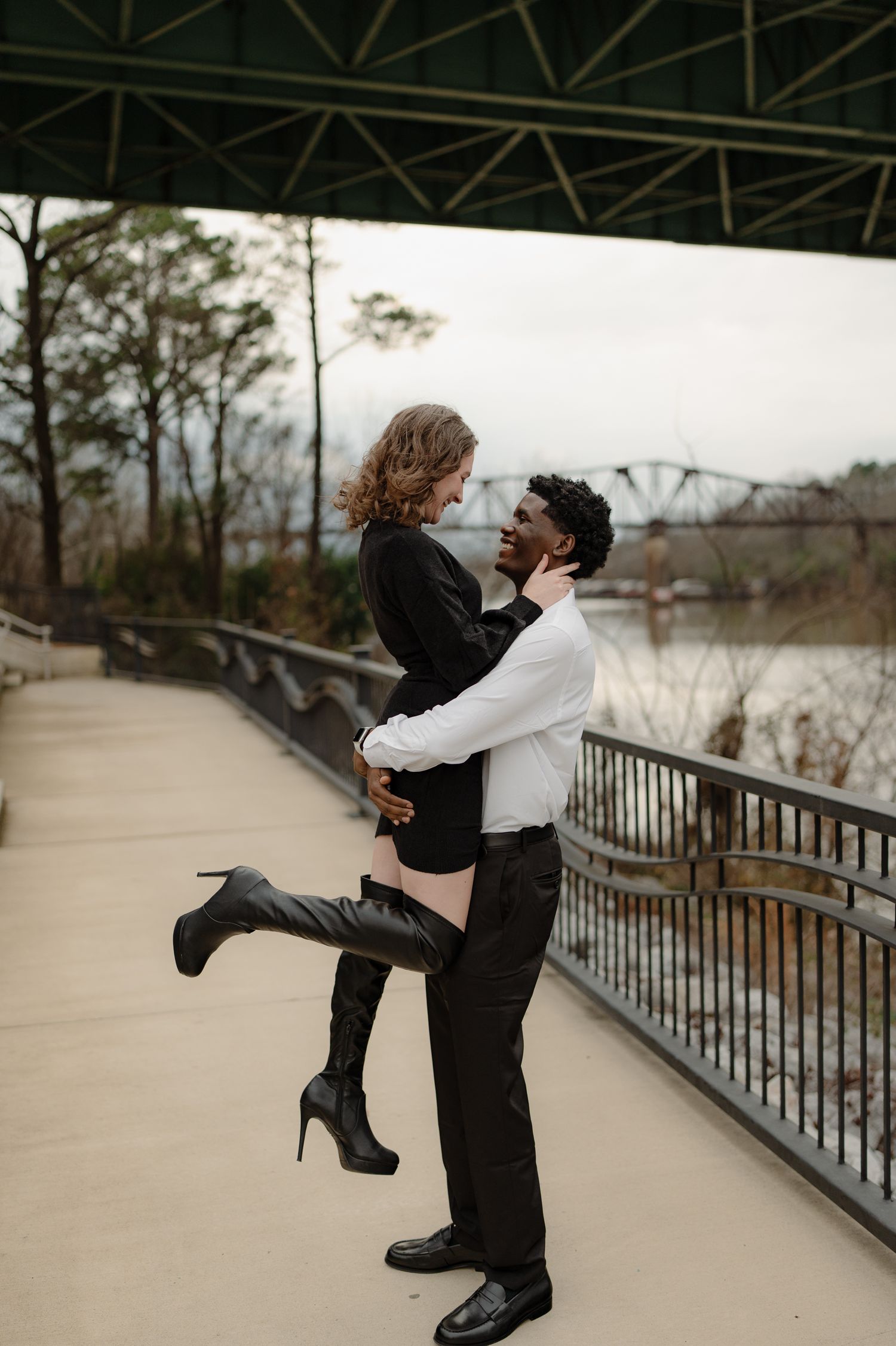 A man lifts a woman in a black dress and knee-high boots on a scenic riverside walkway.