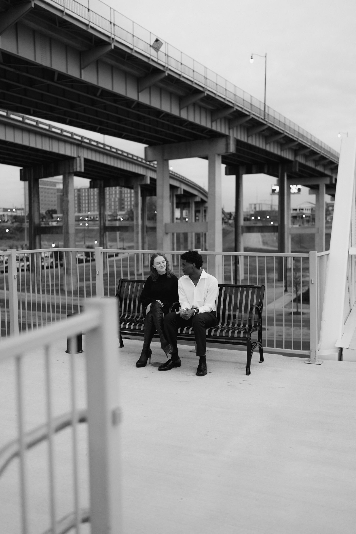 A couple sits closely on a bench beneath a highway bridge in a black and white urban outdoor photo.