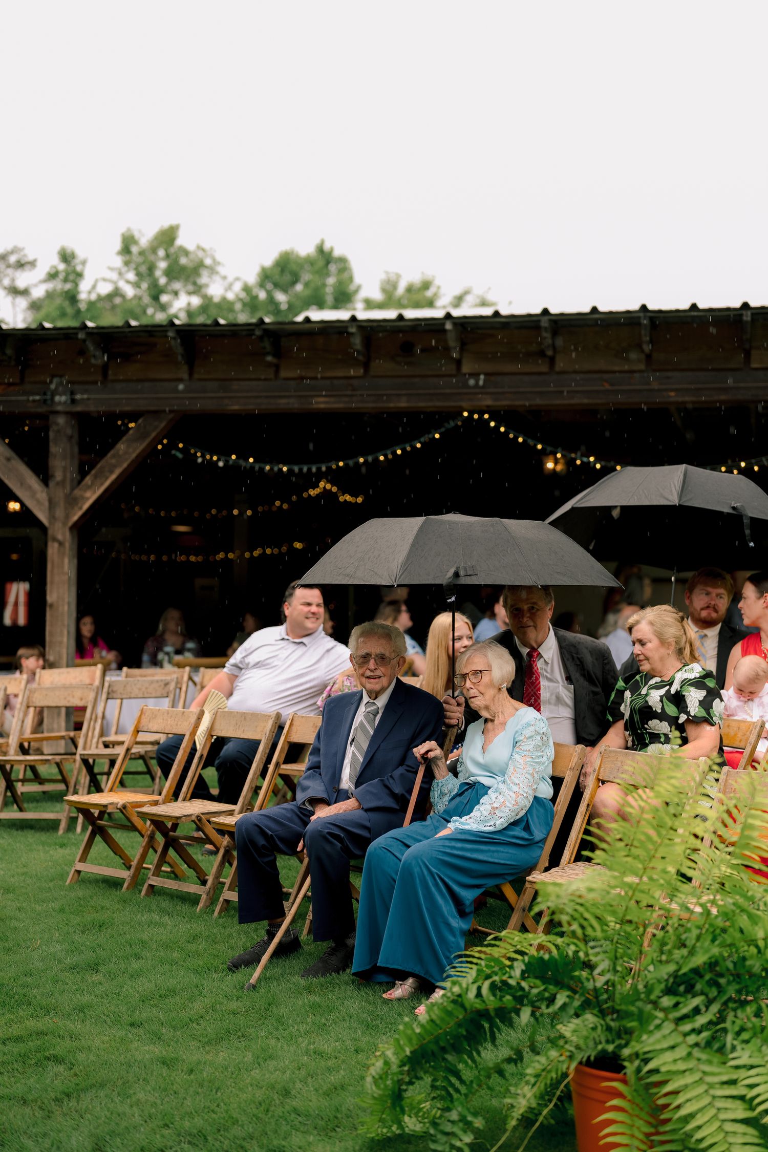 Wedding guests seated on wooden chairs at outdoor ceremony with string lights and umbrellas.
