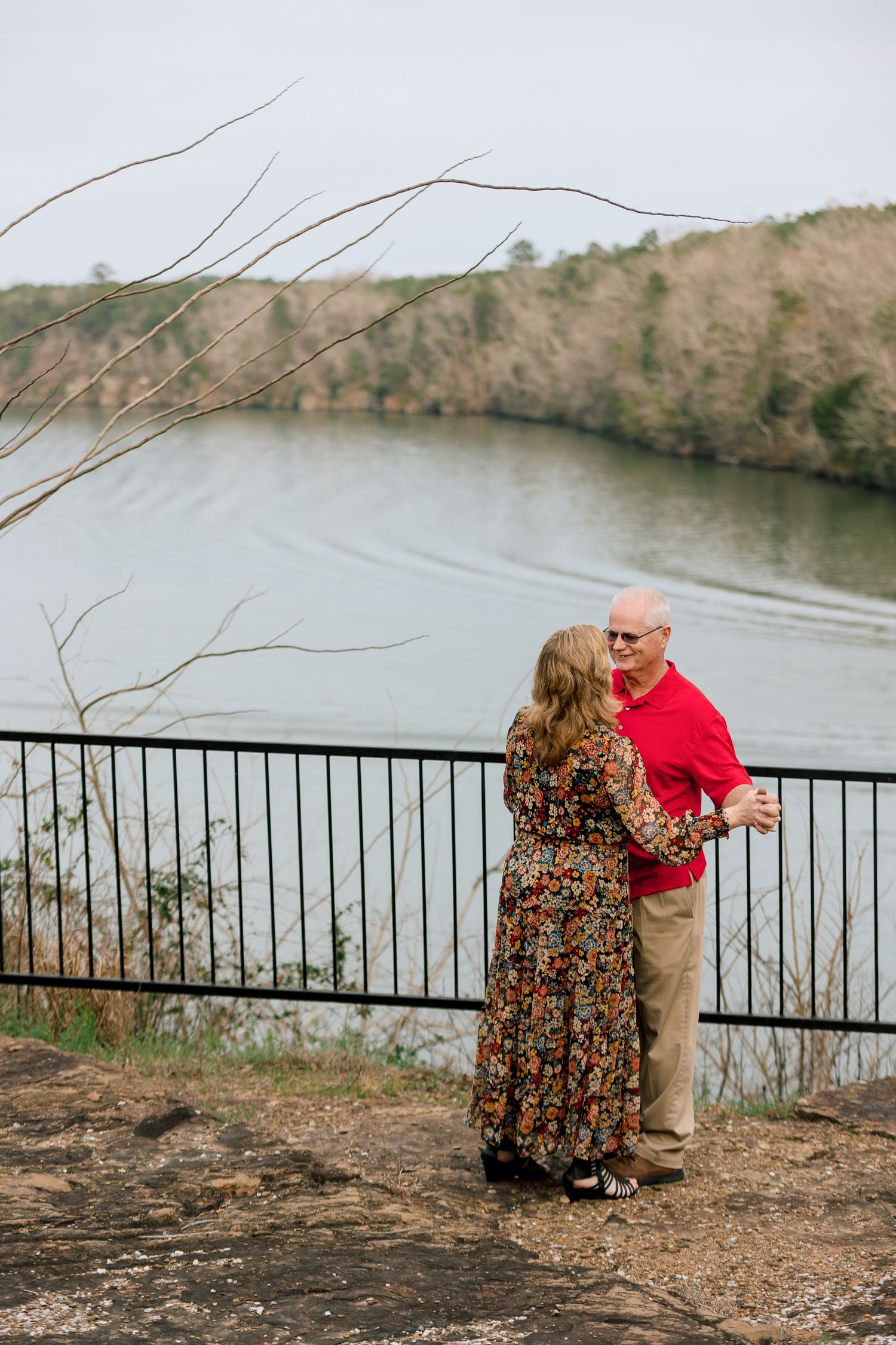 Couple dances and embraces by a railing overlooking a scenic river outdoors.