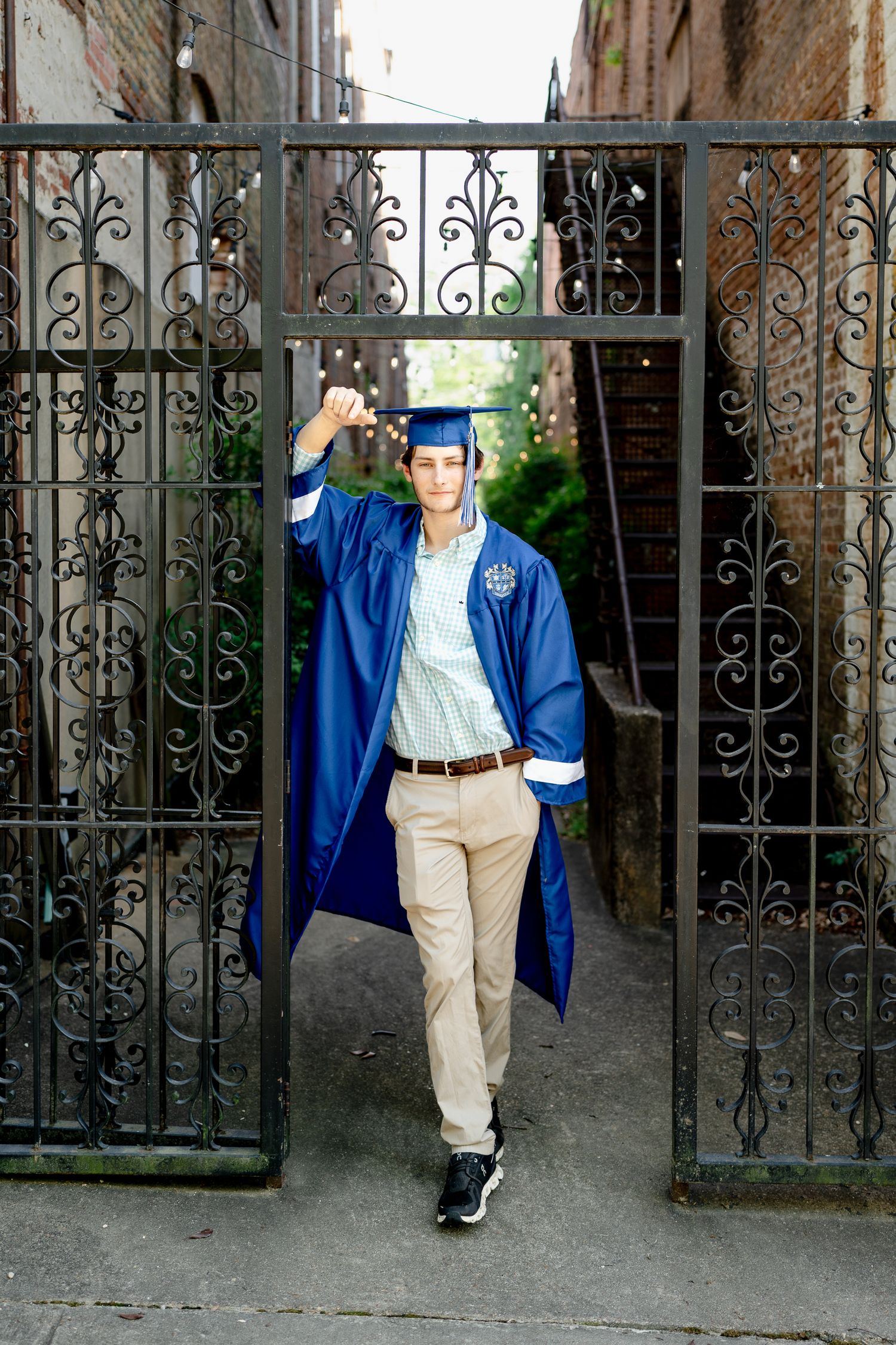 Graduate in blue cap and gown poses by ornate iron gates during graduation photo session.