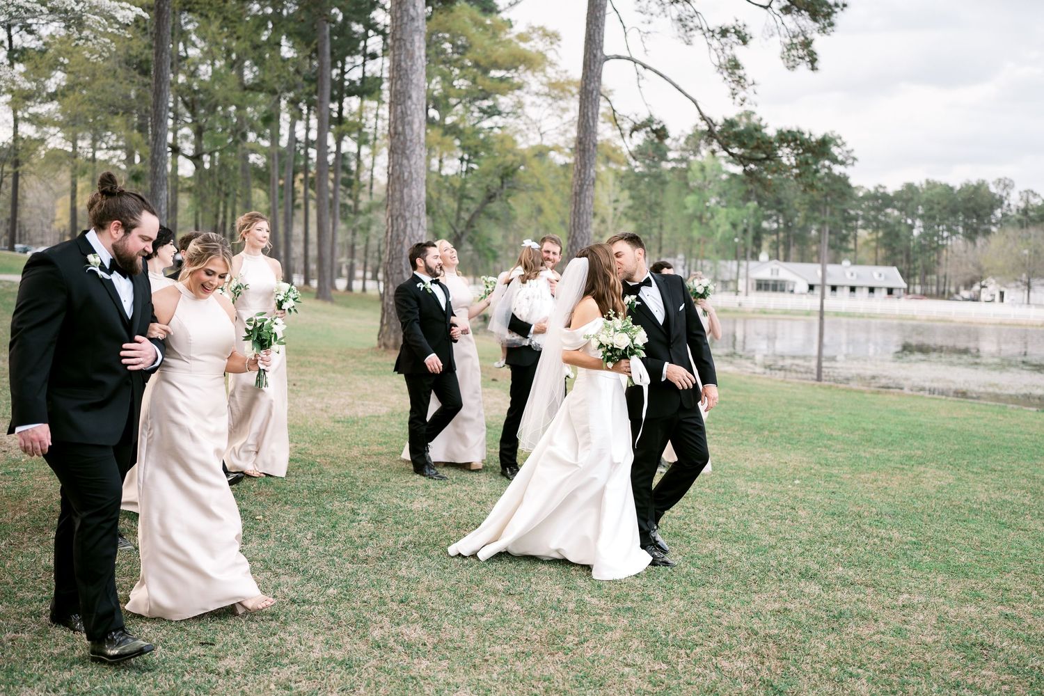 Wedding party dances and celebrates outdoors on green grass with pine trees.