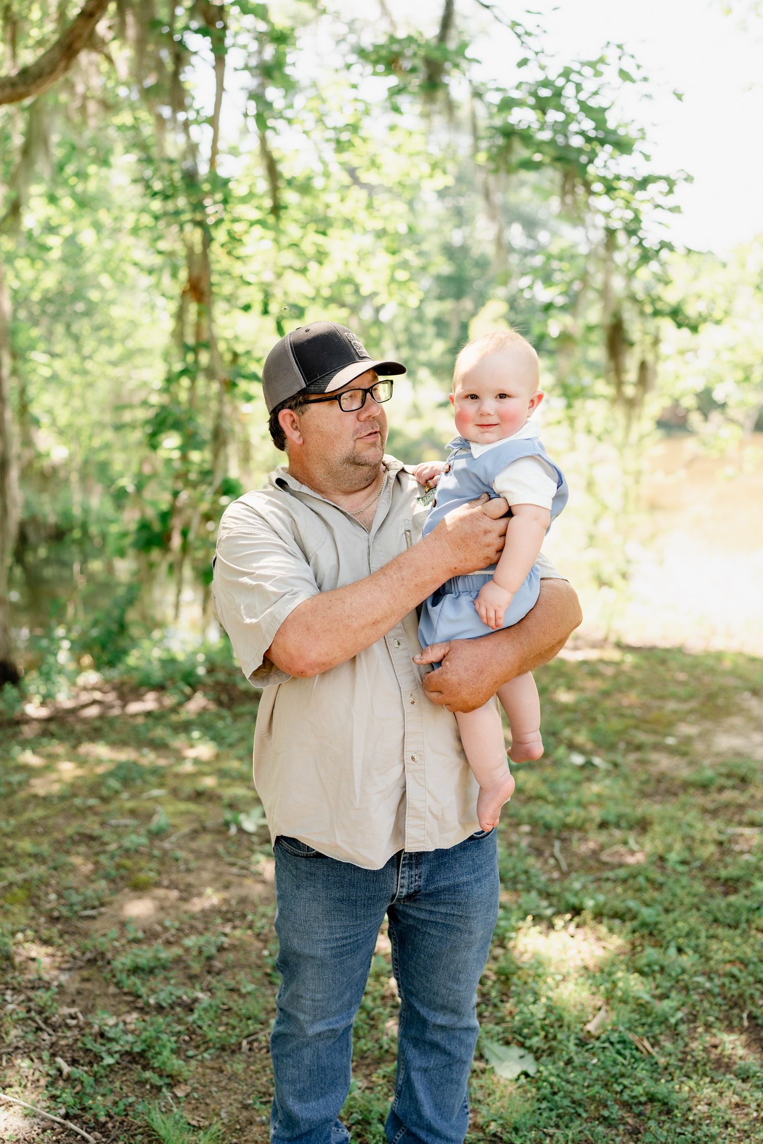 A casual outdoor family portrait showing a man in a baseball cap holding a baby in a wooded setting.