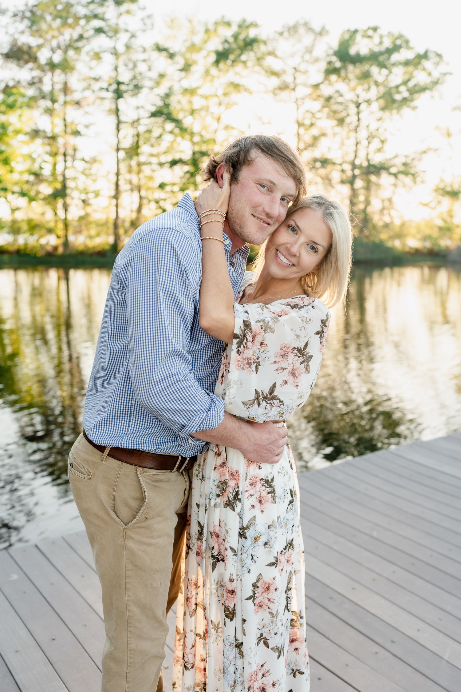 A couple embraces while standing on a wooden dock with a lake and pine trees in the background.