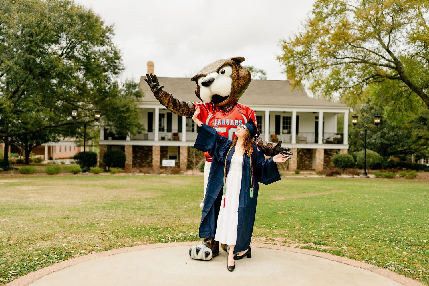 A playful mascot in graduation attire poses on a pathway with a stately building in the background.
