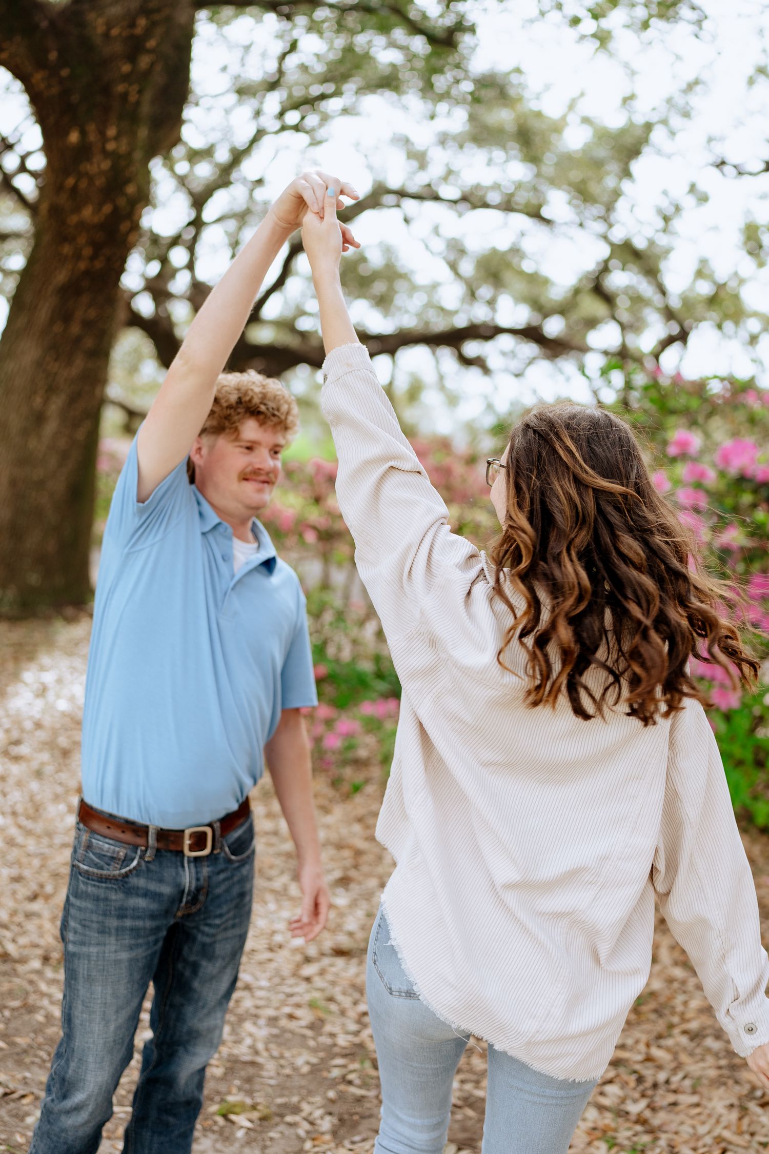 Two people dancing joyfully under oak trees with pink flowers in the background.