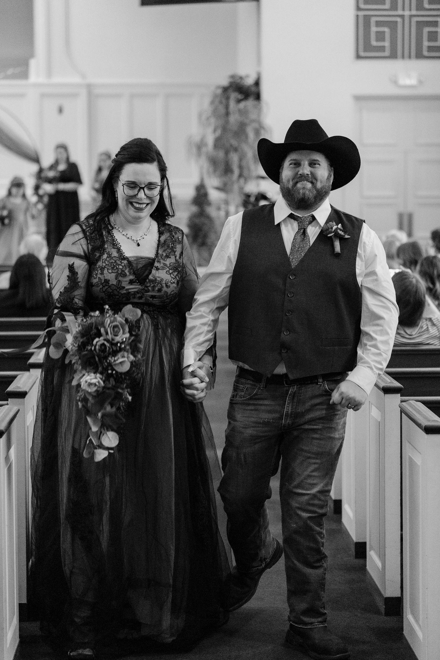Wedding party members in black dresses and vests walk down church aisle during ceremony procession.