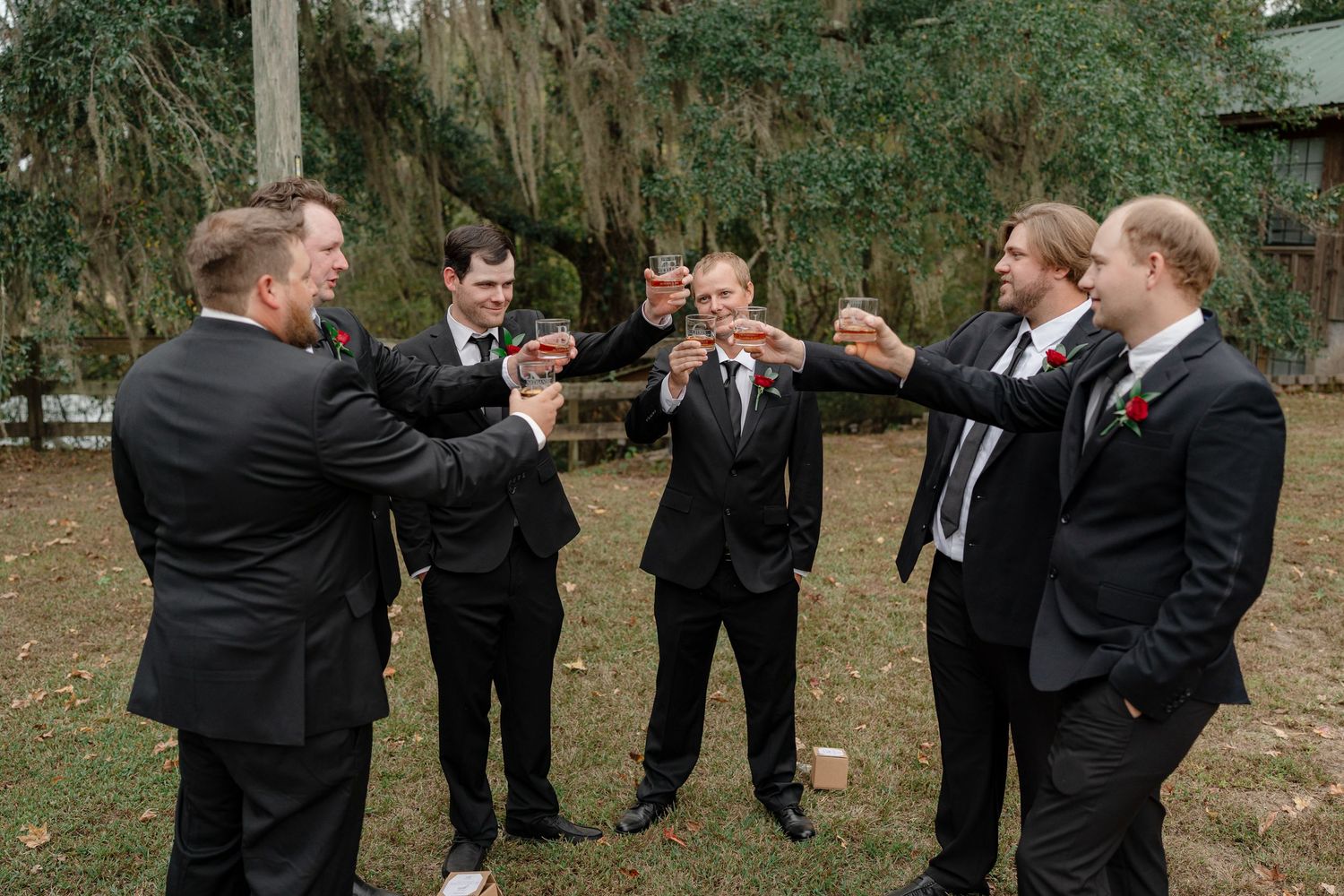 Groomsmen in black suits gather together for casual conversation during outdoor wedding ceremony.