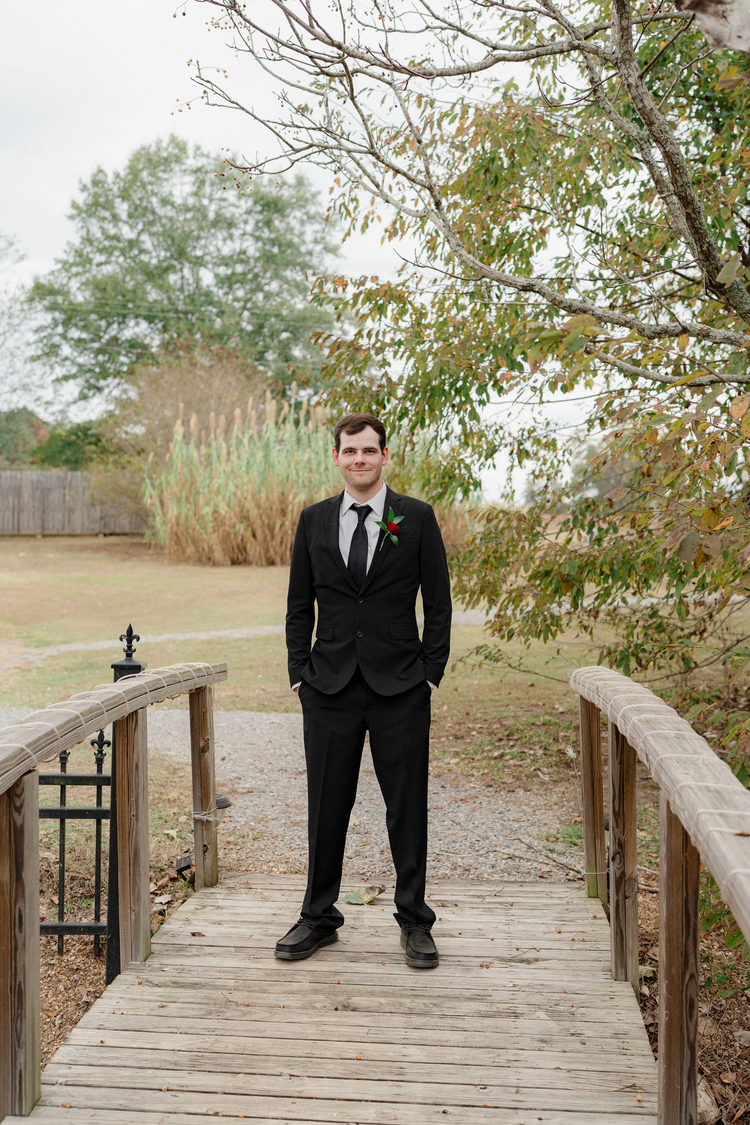 A person in a black suit stands on a wooden bridge walkway surrounded by autumn trees and ornamental grasses.