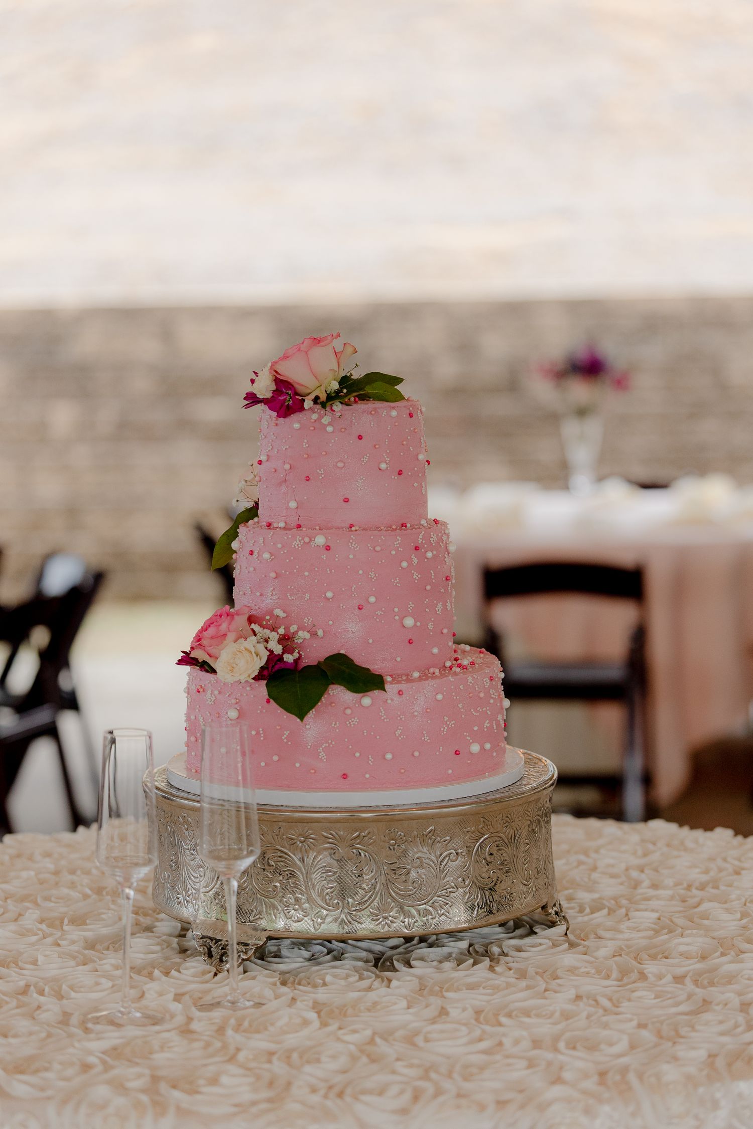Elegant three-tier pink wedding cake adorned with pearls and fresh flowers displayed on ornate silver cake stand.