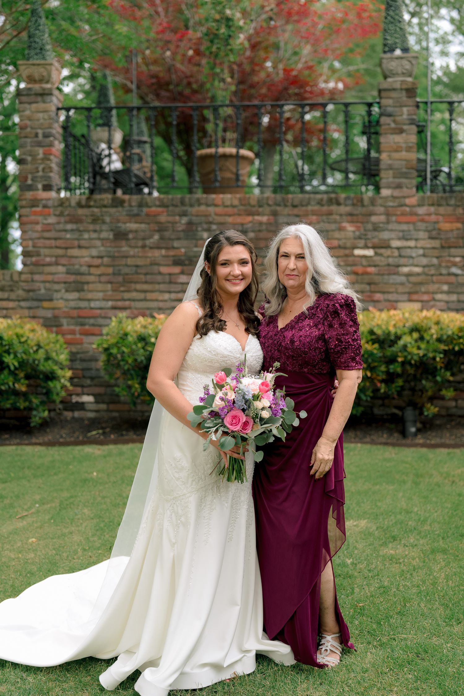 Bride in fitted lace gown holding pink bouquet posing with mother in burgundy dress at garden venue.