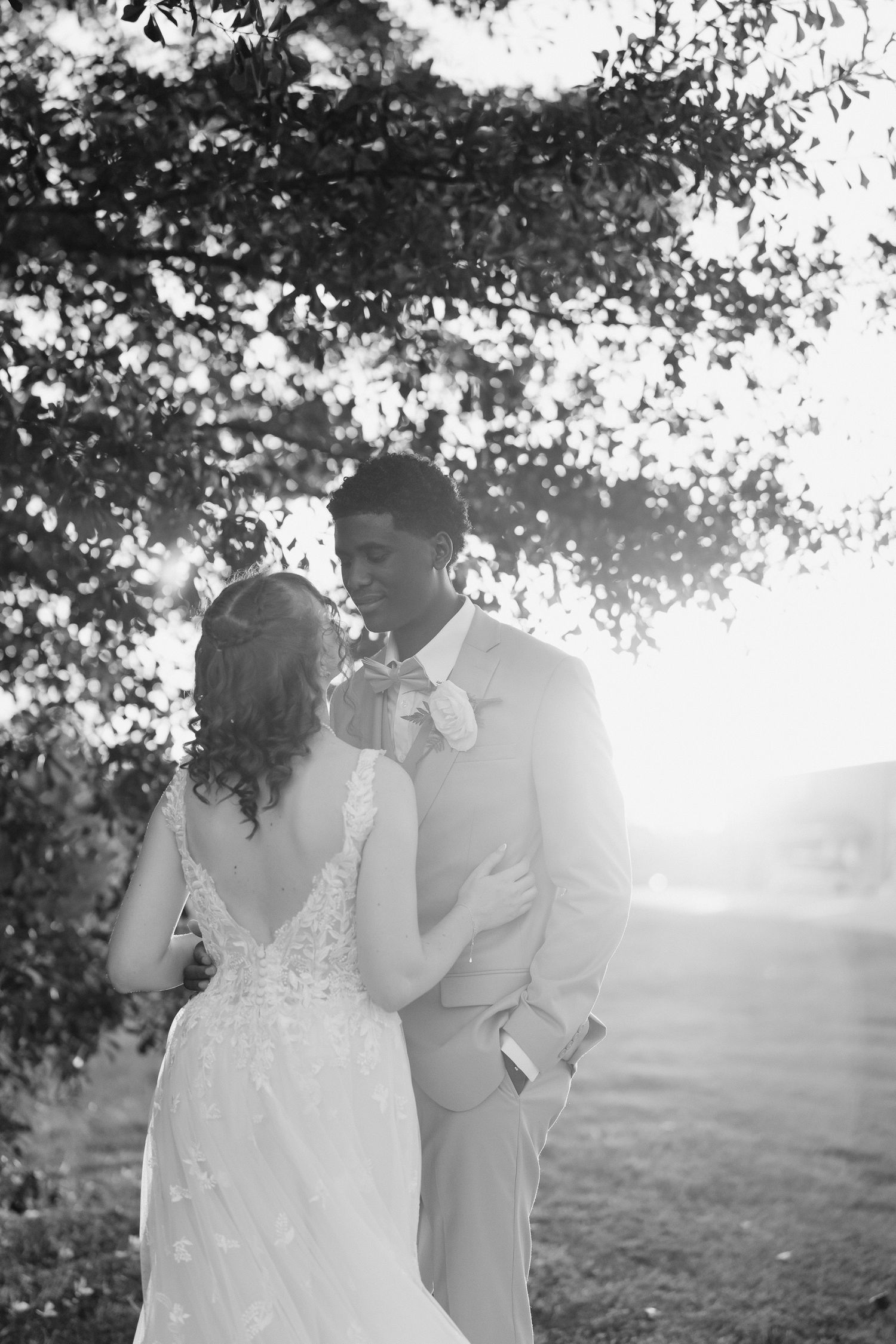 Black and white photo of bride and groom sharing romantic moment under sunlit tree at outdoor wedding.