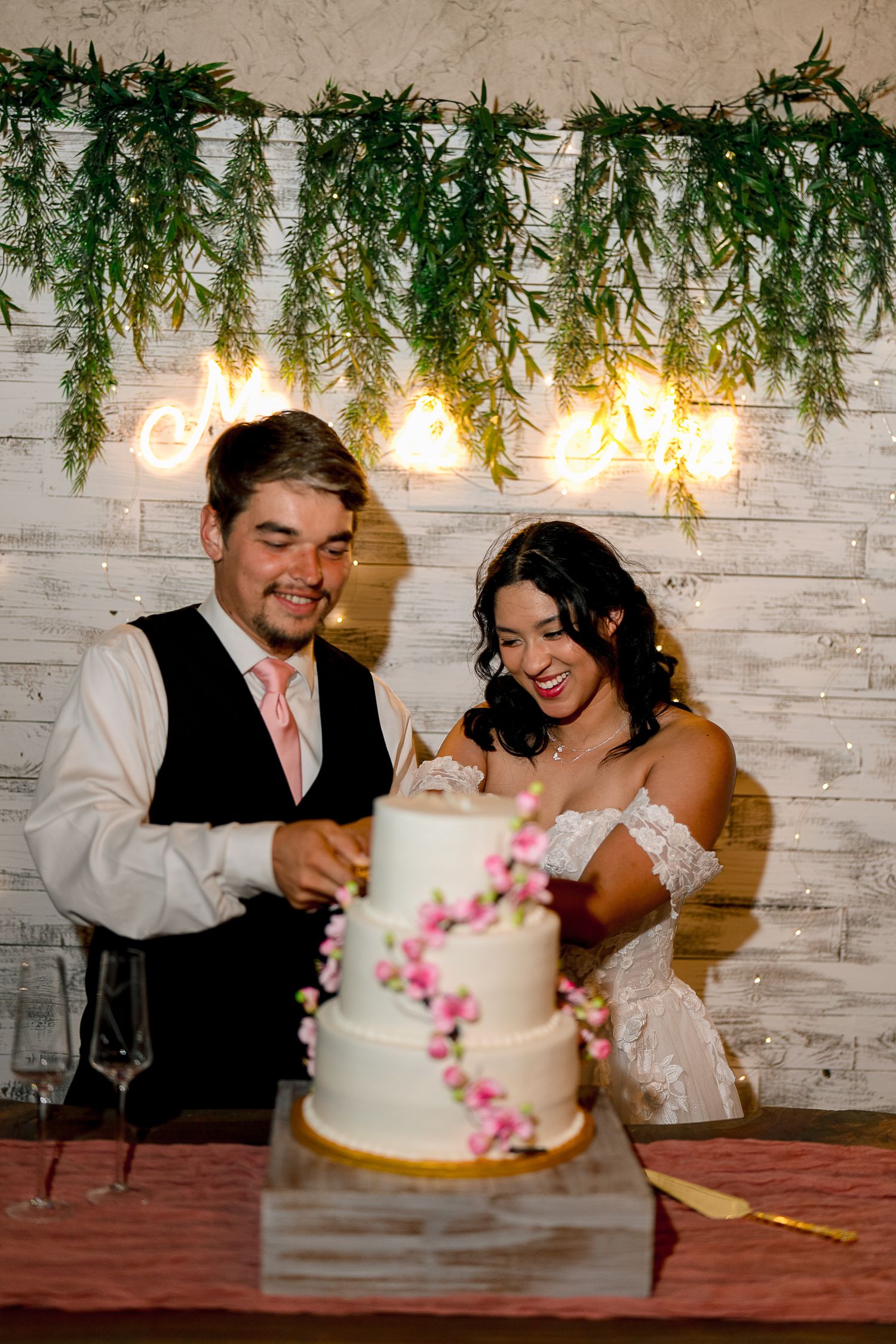 Smiling newlyweds cut their elegant three-tier white wedding cake decorated with pink floral accents.