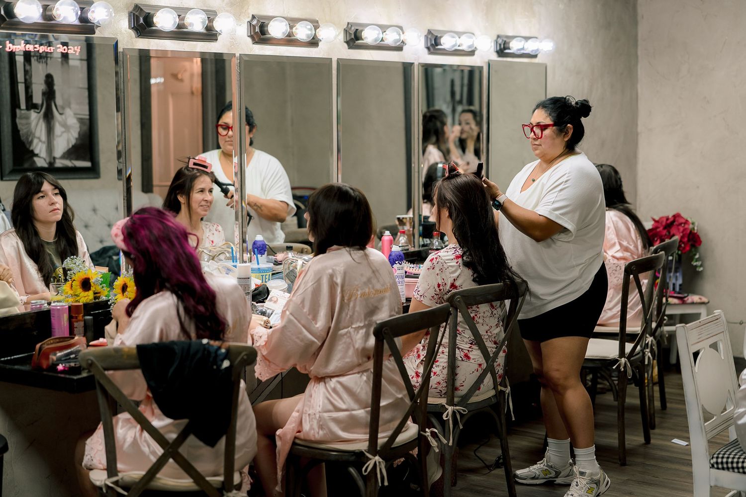 Bridesmaids in robes getting hair and makeup done in a vanity room with lit mirrors.