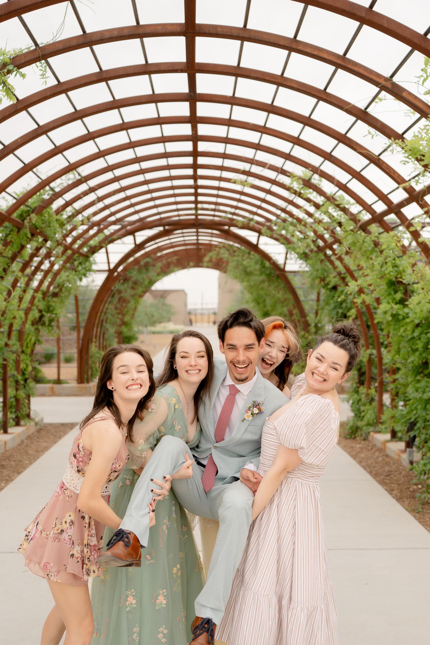 Wedding party members laugh together under a rustic wooden pergola with greenery.
