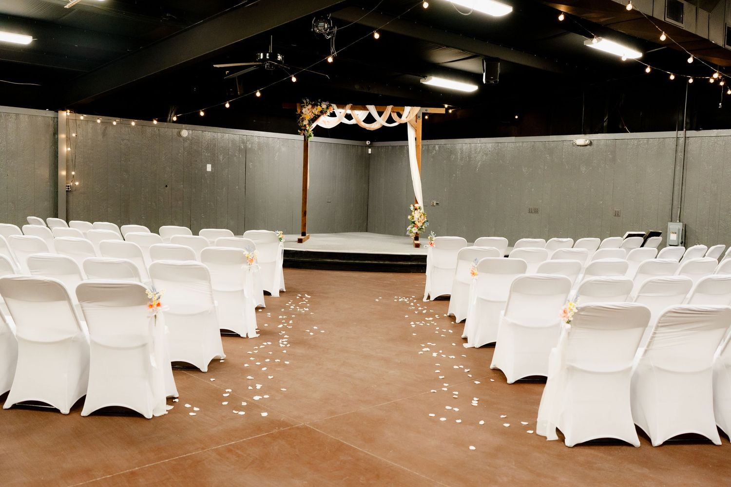 Indoor event space with rows of white covered chairs set up for a ceremony under exposed ceiling beams and string lights.