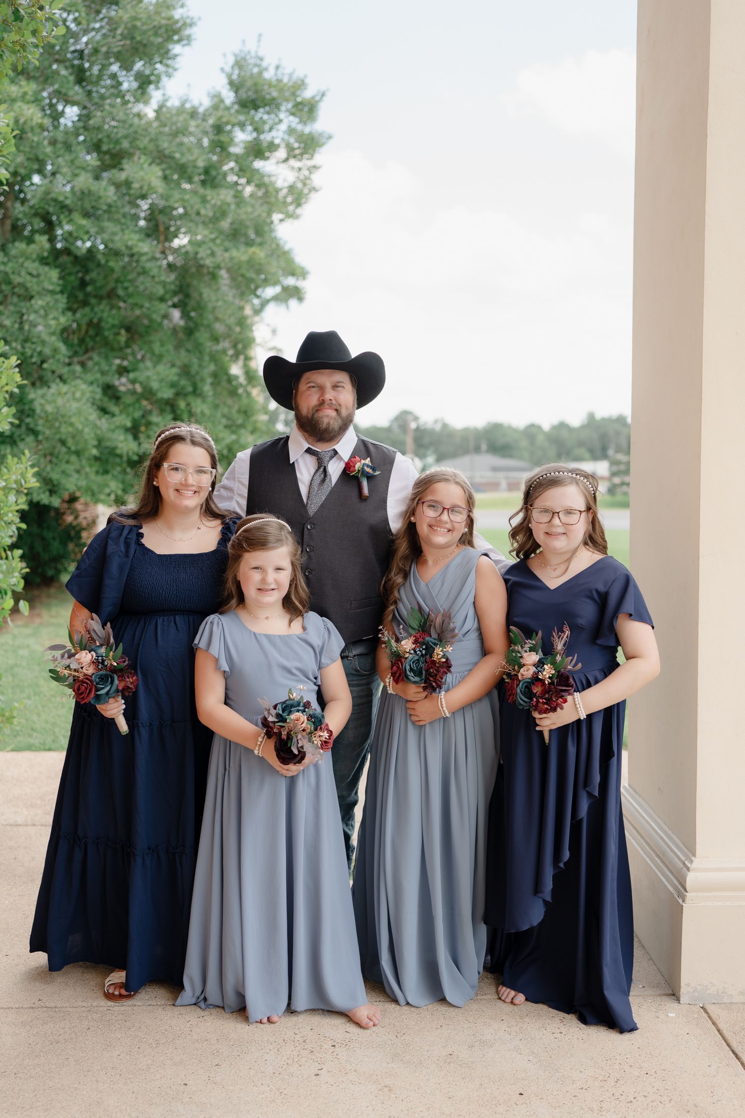 Wedding party in navy blue and gray dresses poses together under white columns outdoors.