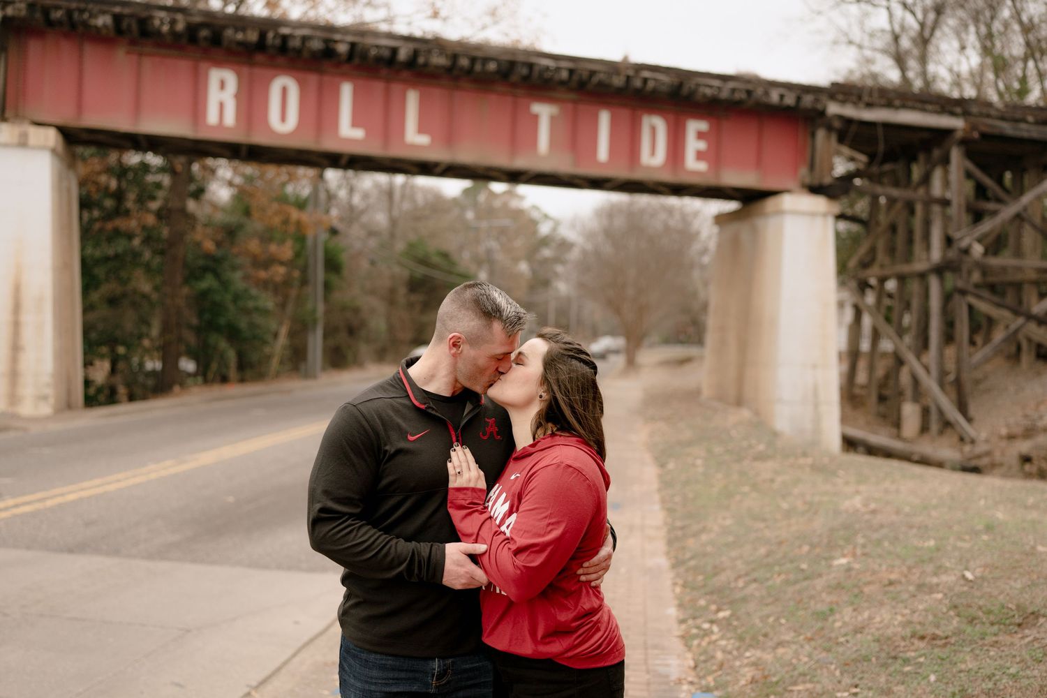 A couple shares an intimate moment beneath a Roll Tide sign on a bridge.