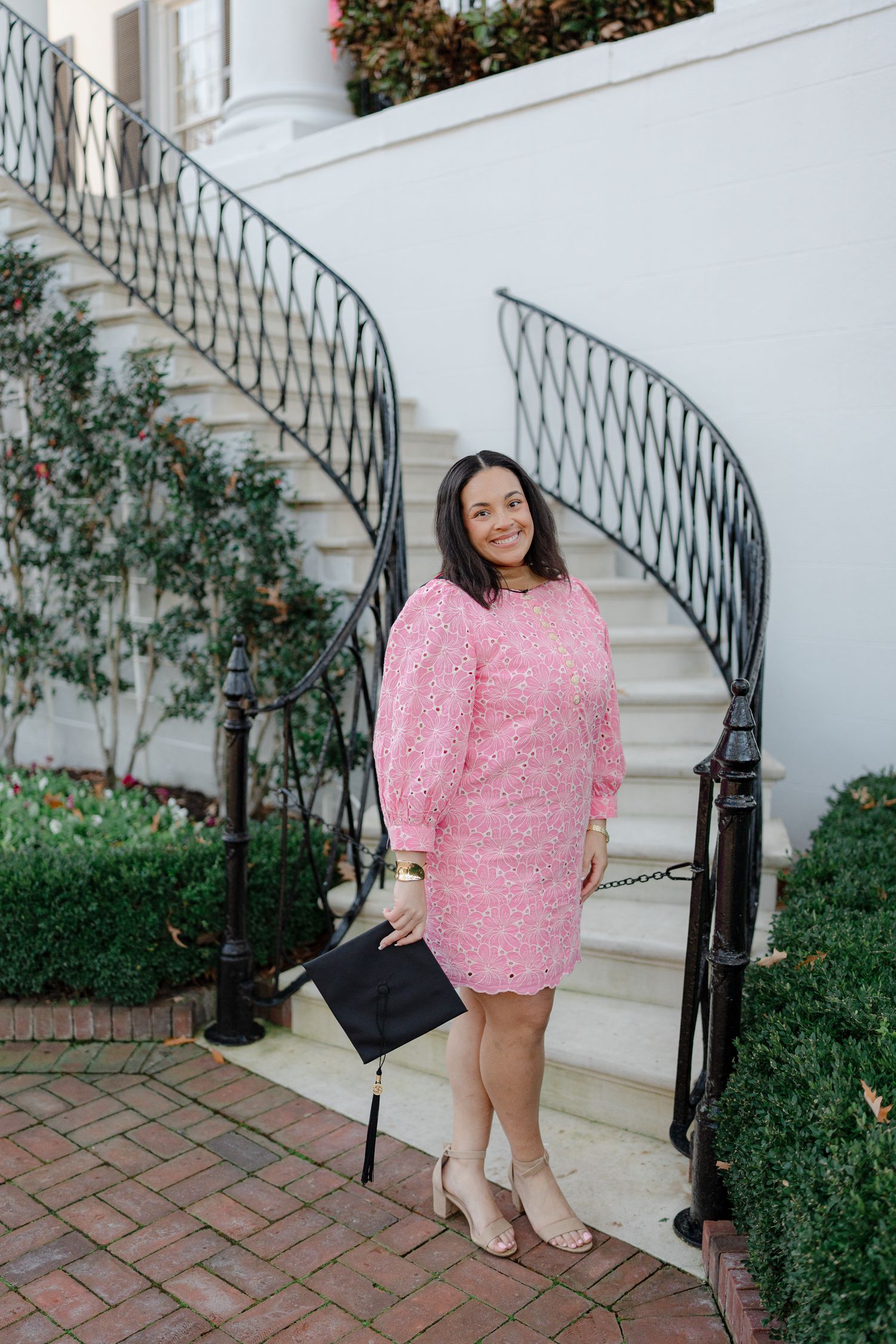 A graduate in pink lace dress stands by an elegant curved staircase with wrought iron railings.