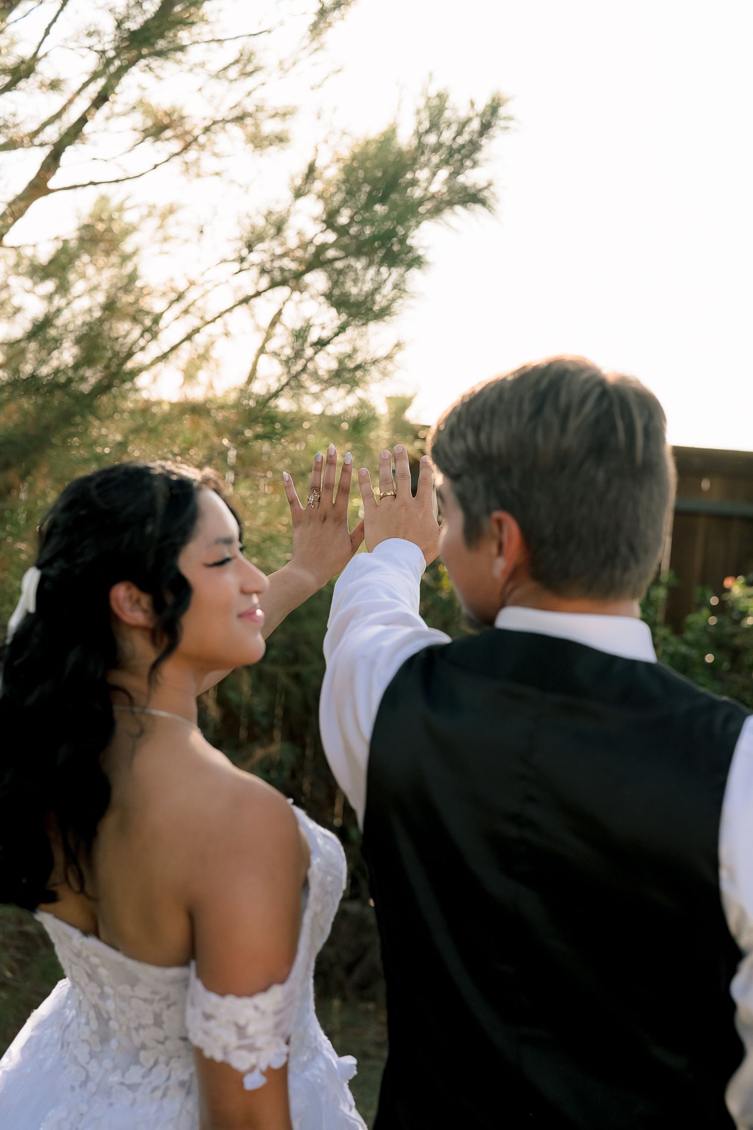 Bride in lace off-shoulder gown smiles as groom gently touches her face during golden hour outdoors.