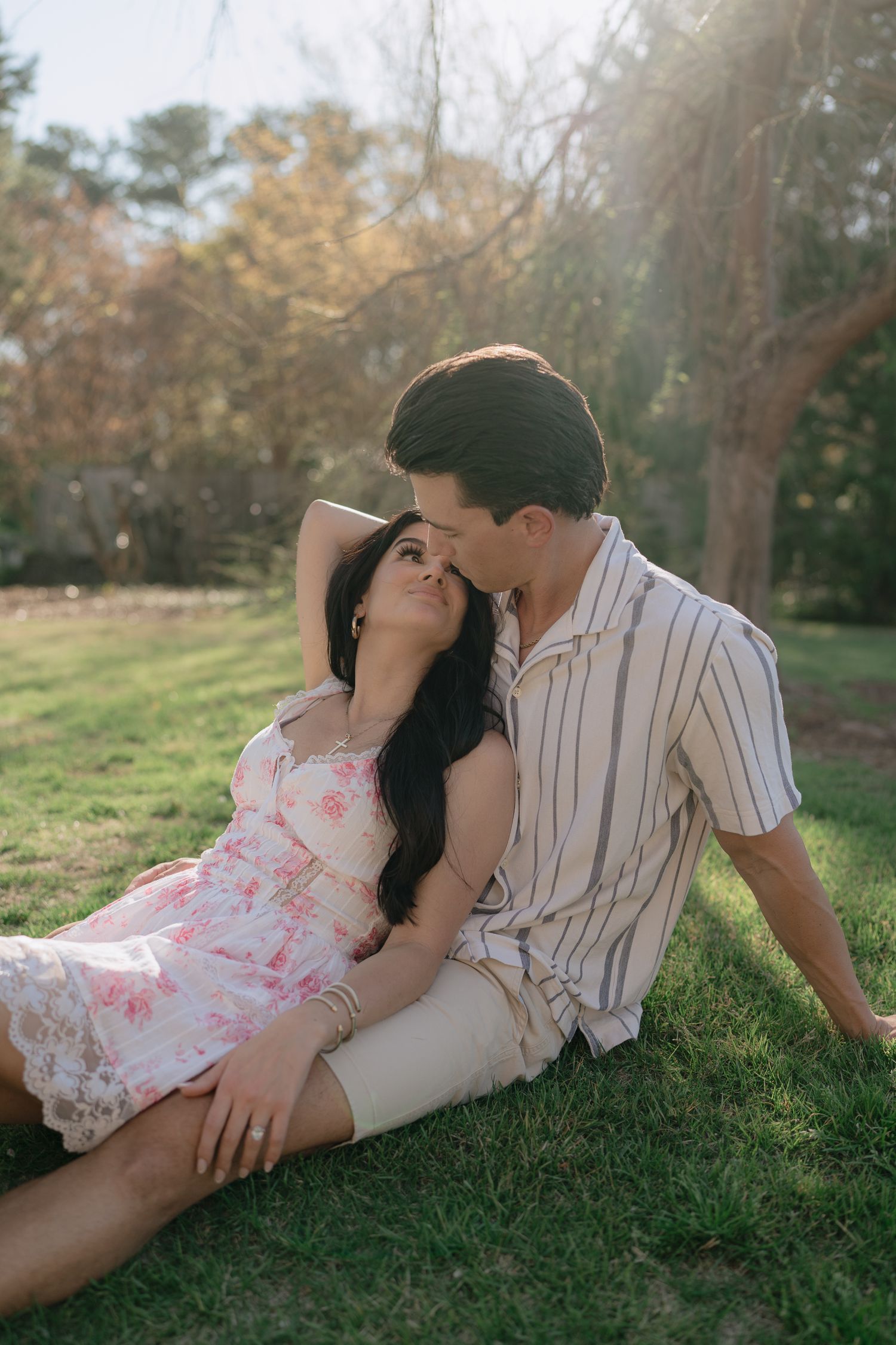 A couple sits together on green grass in golden sunlight, man gently kissing womans temple.