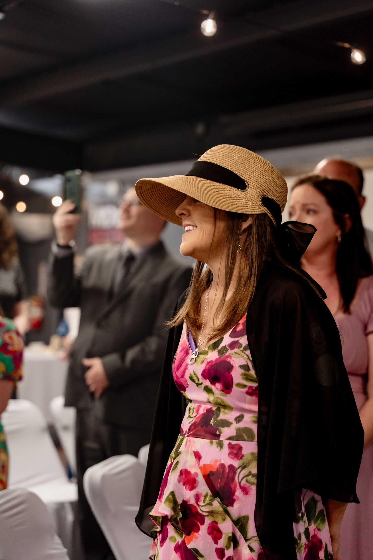 A guest in a floral dress and wide-brimmed hat enjoying a celebratory event indoors.