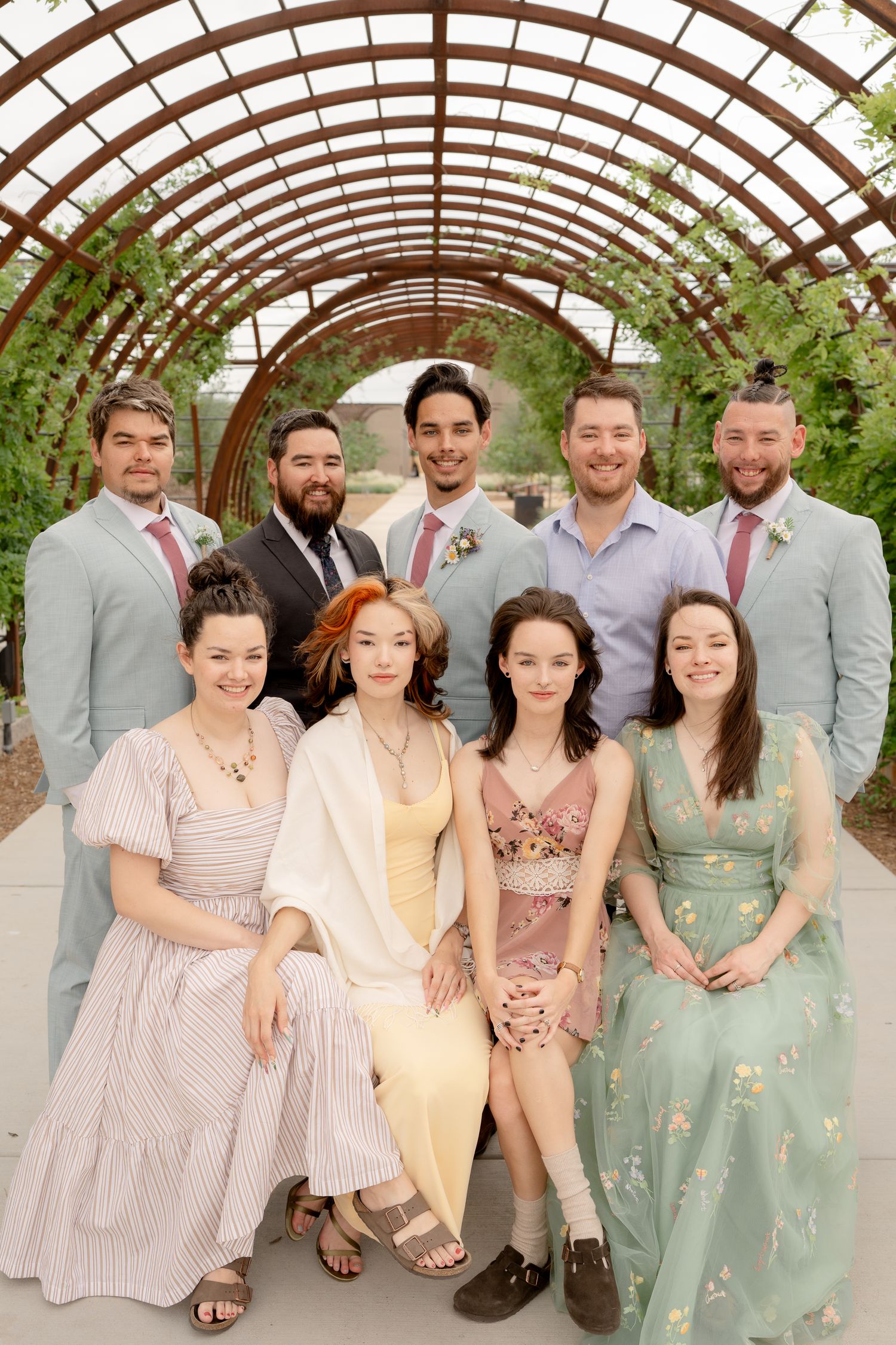 Wedding party in pastel dresses and gray suits pose under a curved greenhouse-style structure with natural lighting.