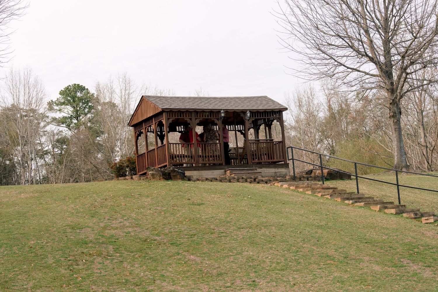 Rustic wooden gazebo pavilion on a green hillside surrounded by bare trees in a park setting.
