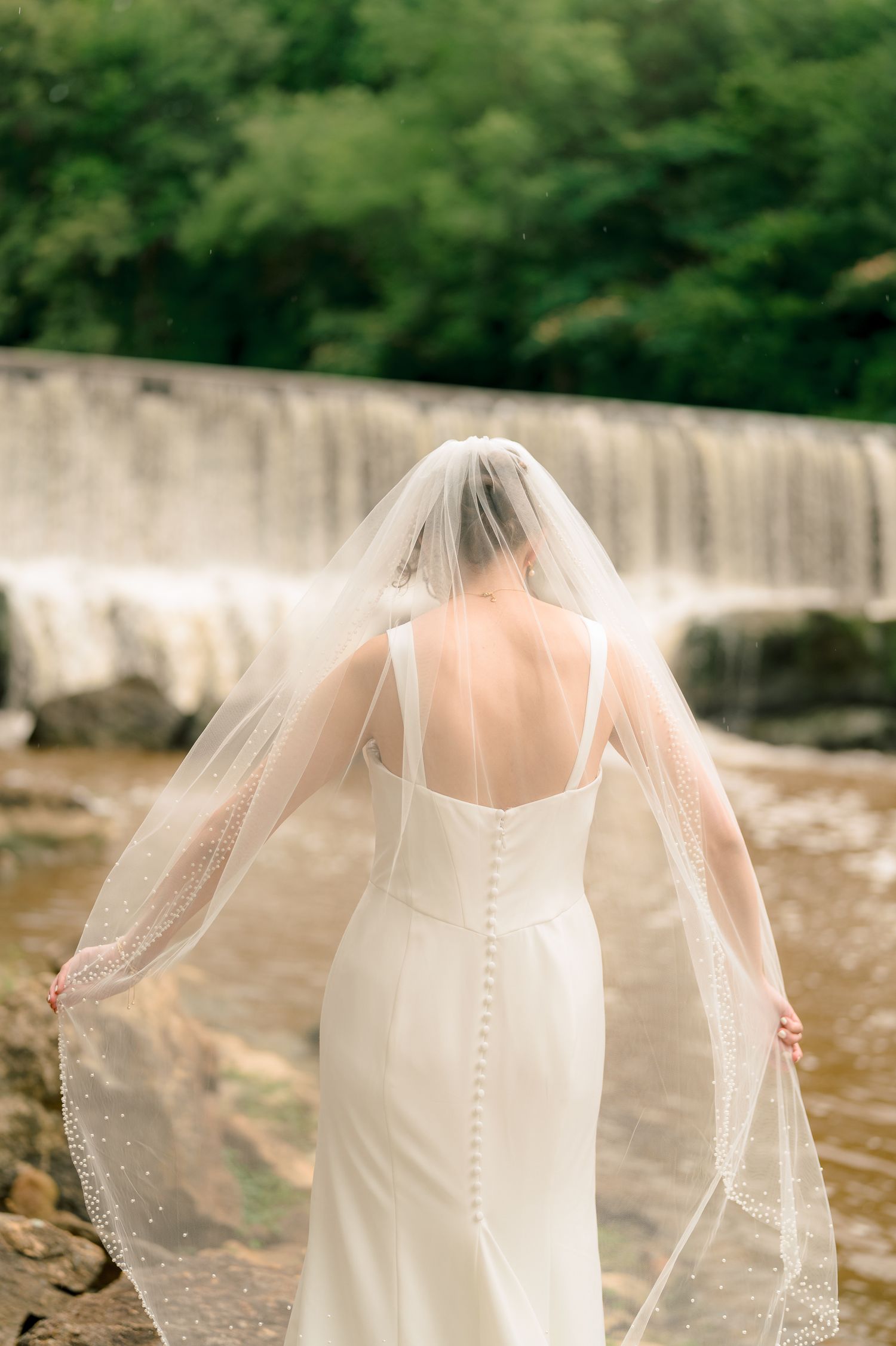 Bride in backless white wedding gown with pearl-button veil standing before scenic waterfall.