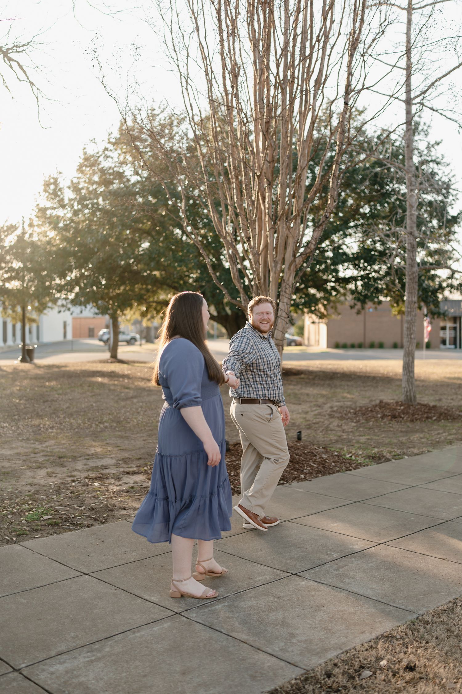 Couple walking and laughing together on a sidewalk path surrounded by bare trees in golden hour light.