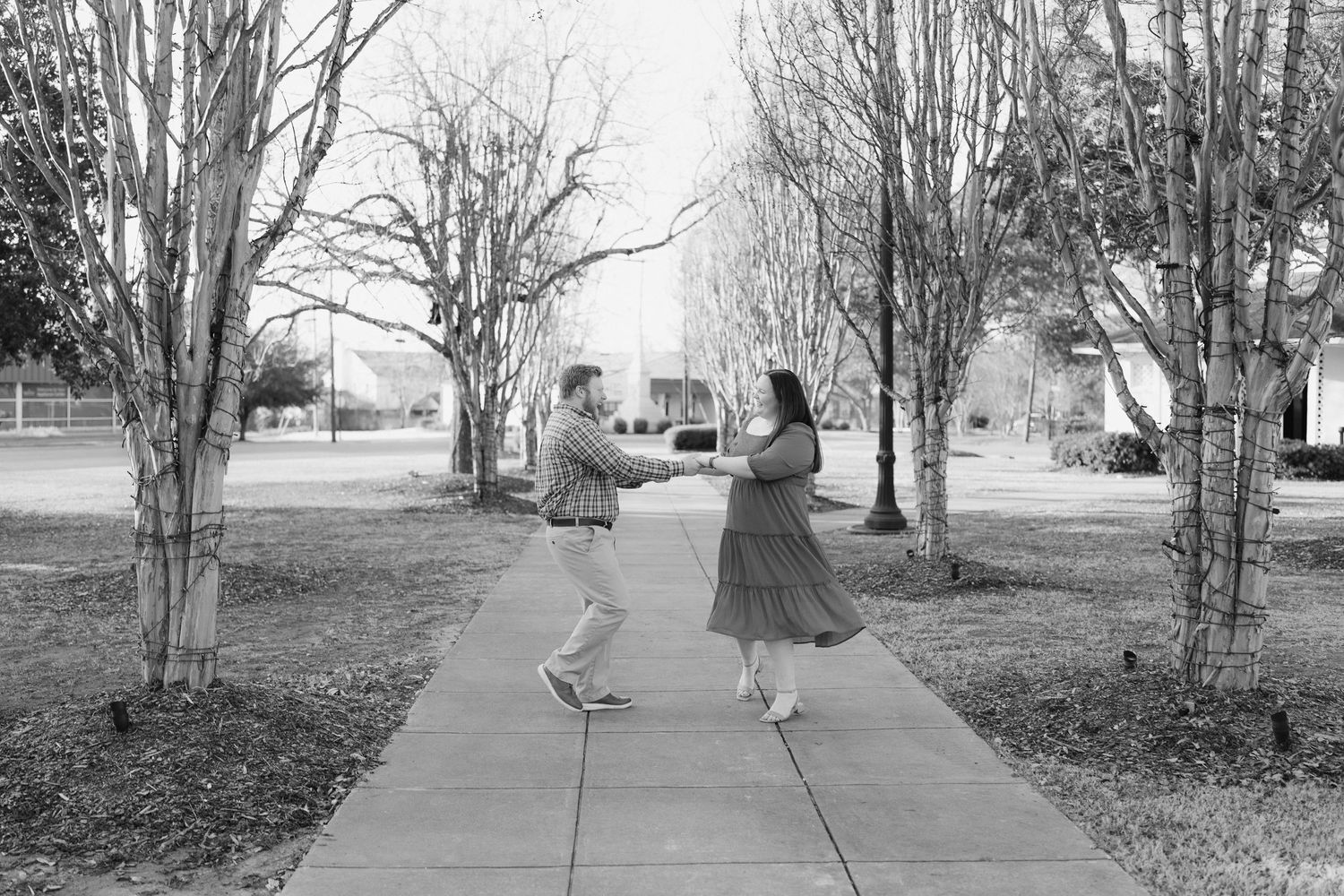 Black and white photo of couple dancing and holding hands on a tree-lined sidewalk in a park.
