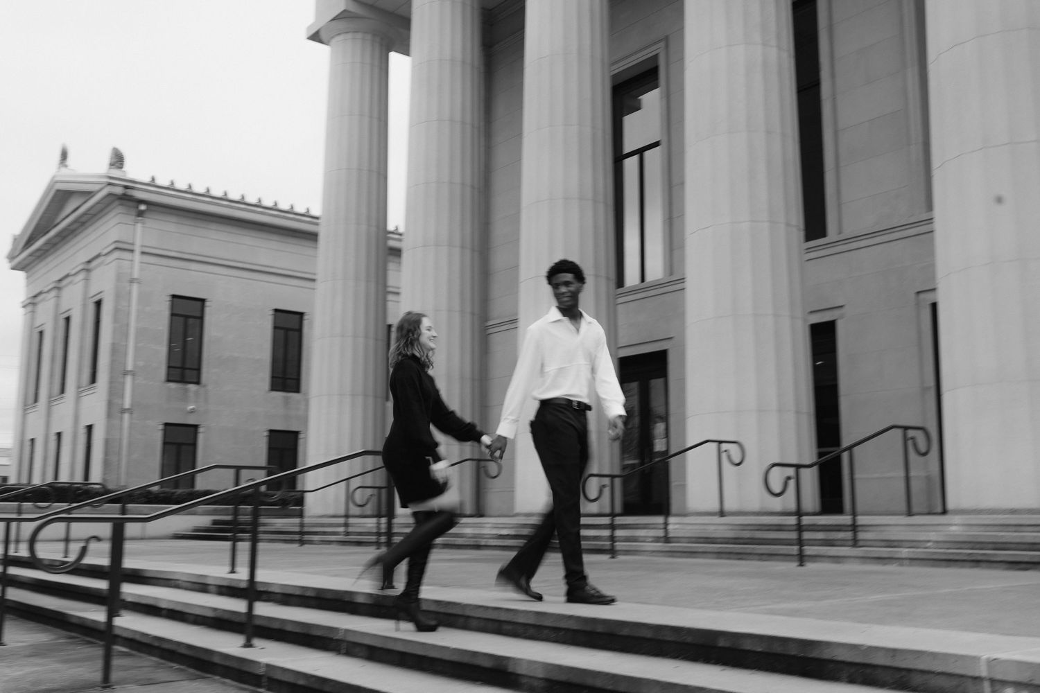 An interracial couple holds hands walking down steps of a grand columned building in black and white.