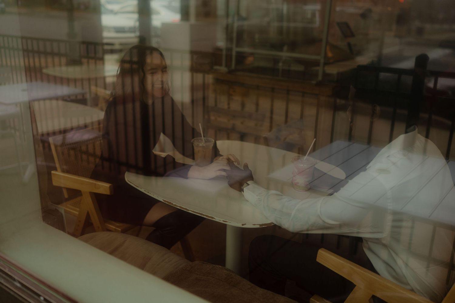 Couple sitting across from each other at a cafe table, viewed through a window reflection.