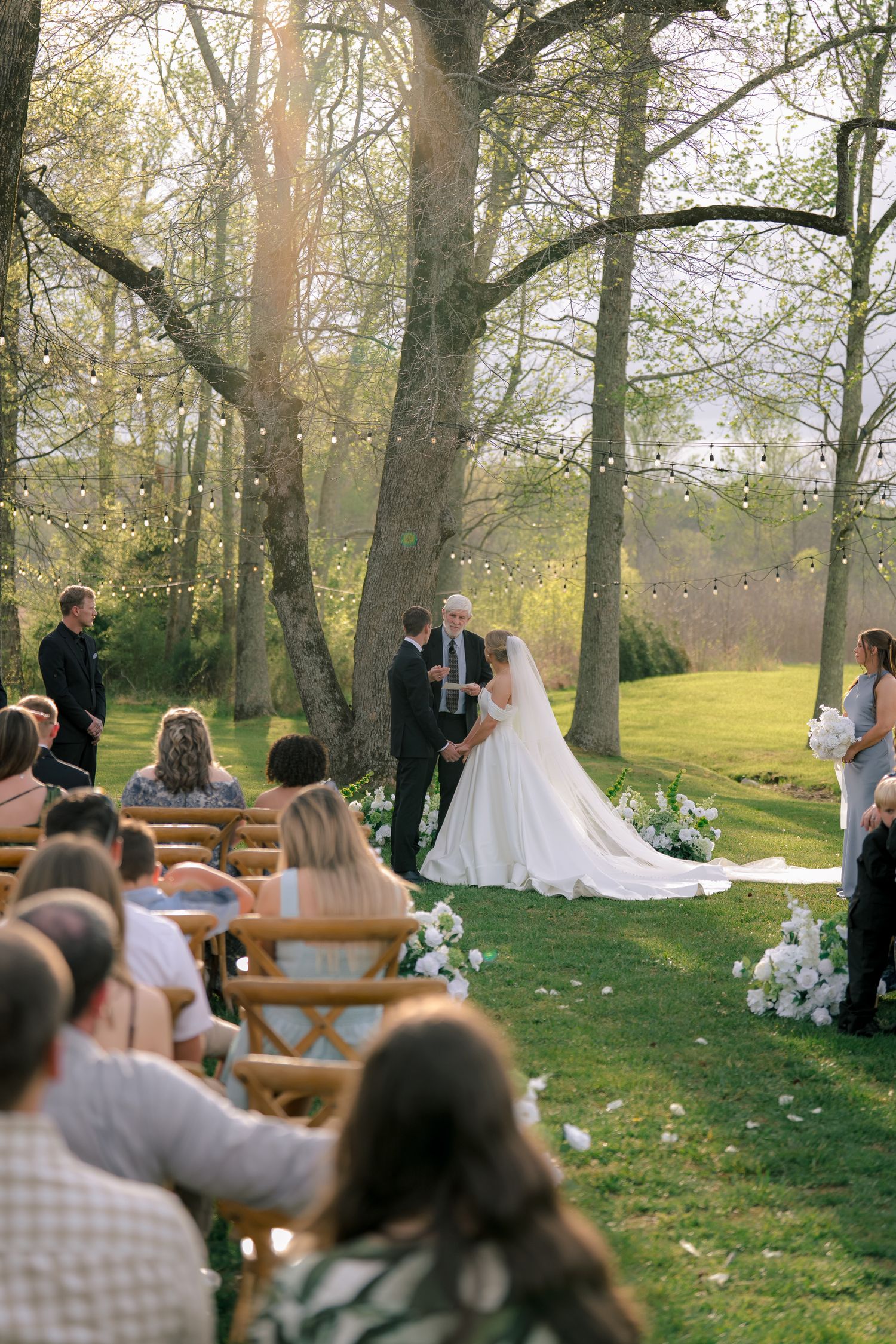 Outdoor wedding ceremony under tall trees with string lights, bride in white gown and long veil at the altar.