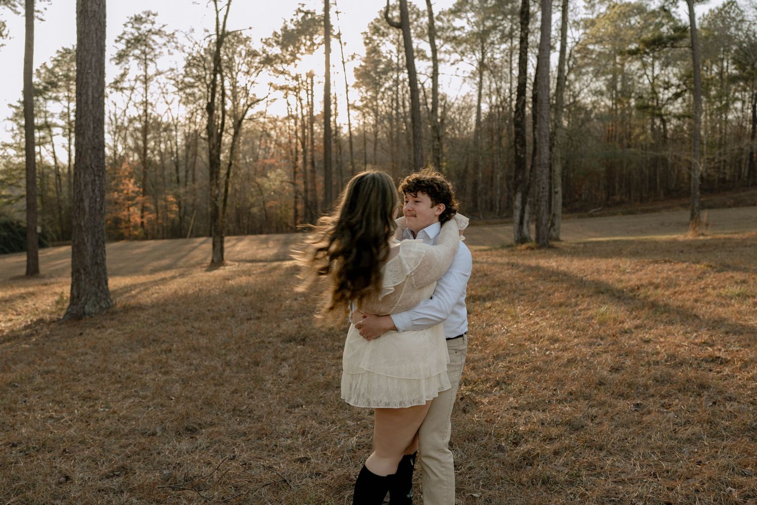 Couple embracing and nearly kissing in a sunlit pine forest during golden hour, woman in white dress and cowboy boots.