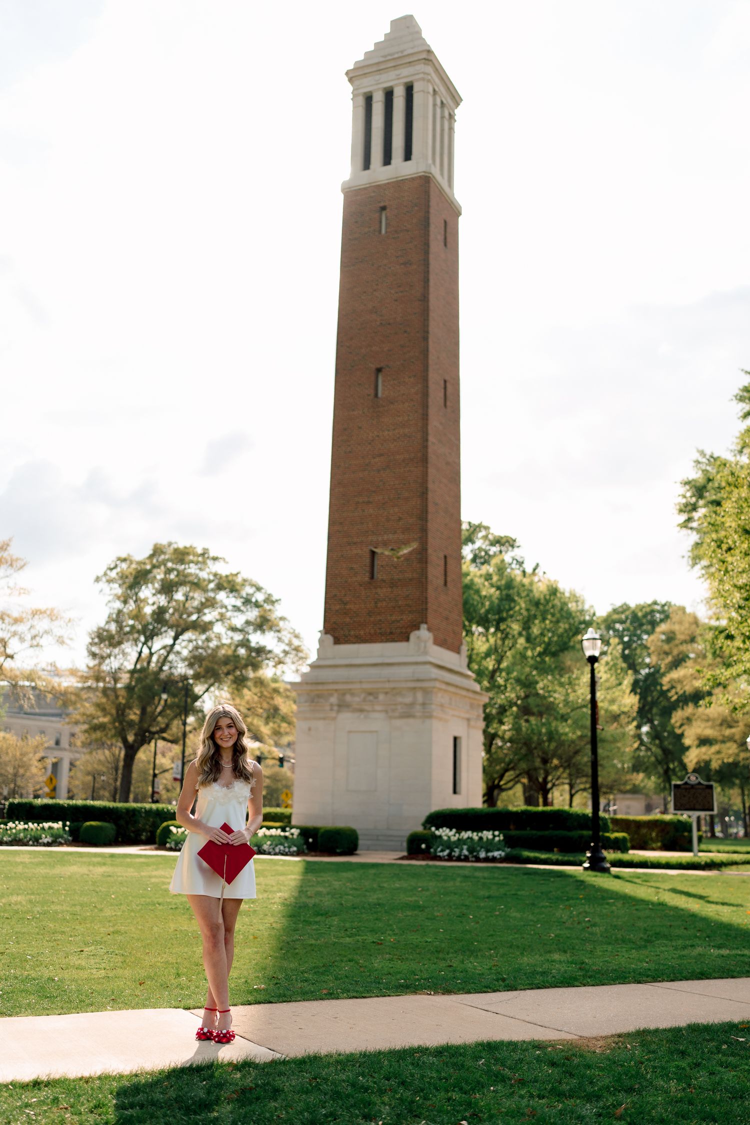 A tall brick bell tower stands on a manicured green lawn with a white dress and red clutch visible in the foreground.