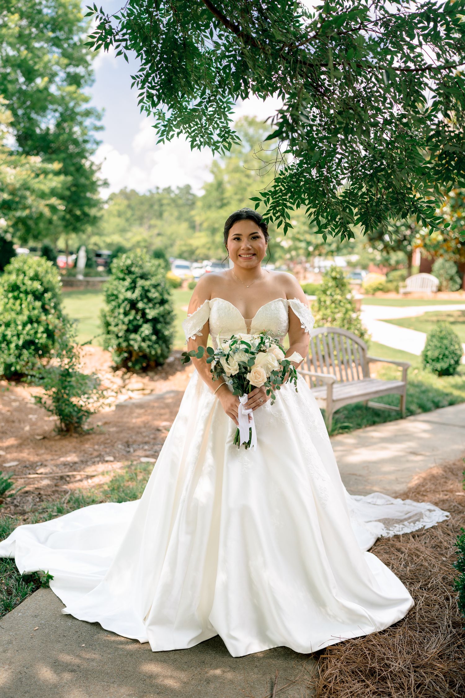 A bride in an elegant off-shoulder white wedding gown holds a bouquet while standing on a garden path under trees.