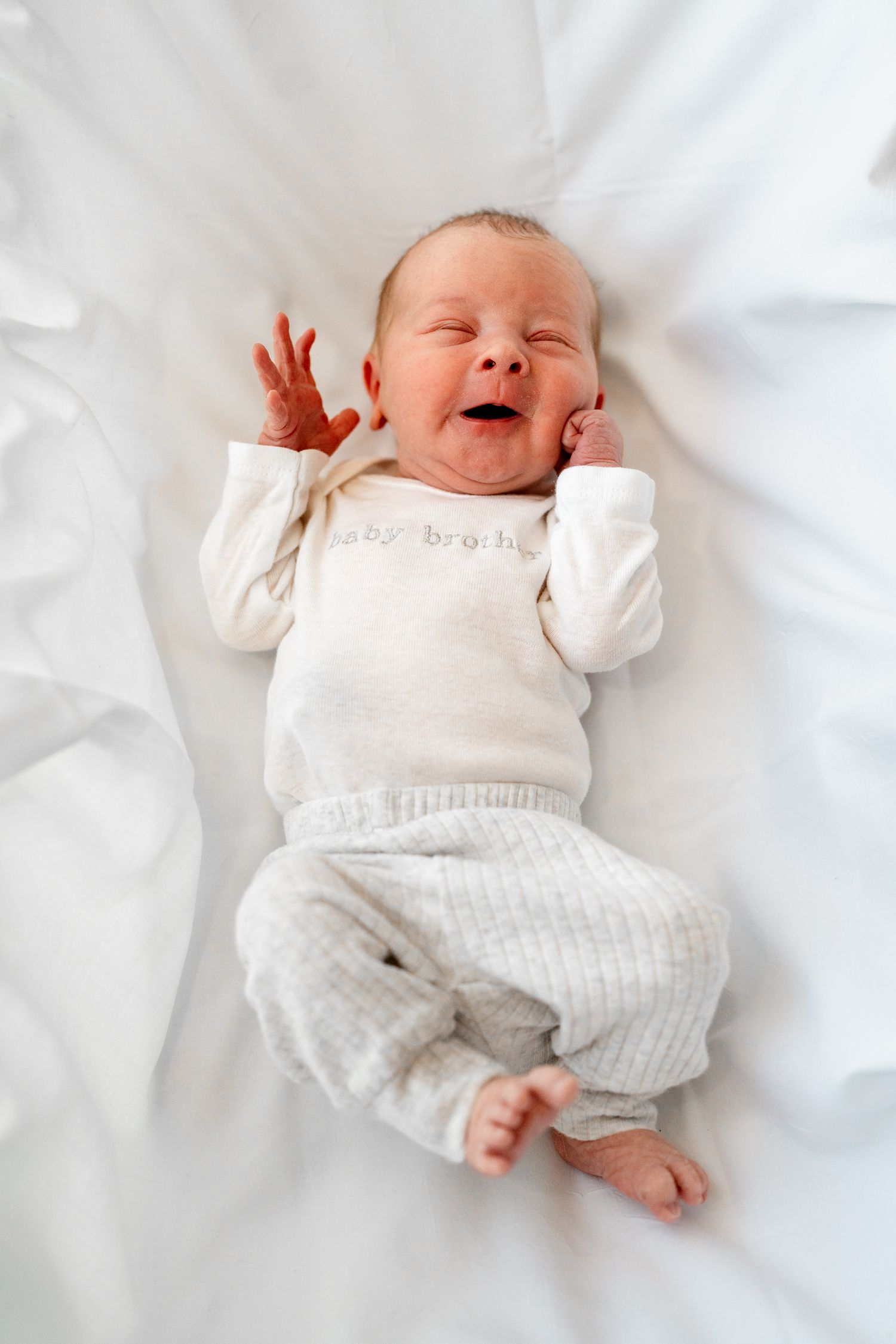 Baby in white knit outfit expressing contentment on white bedding.