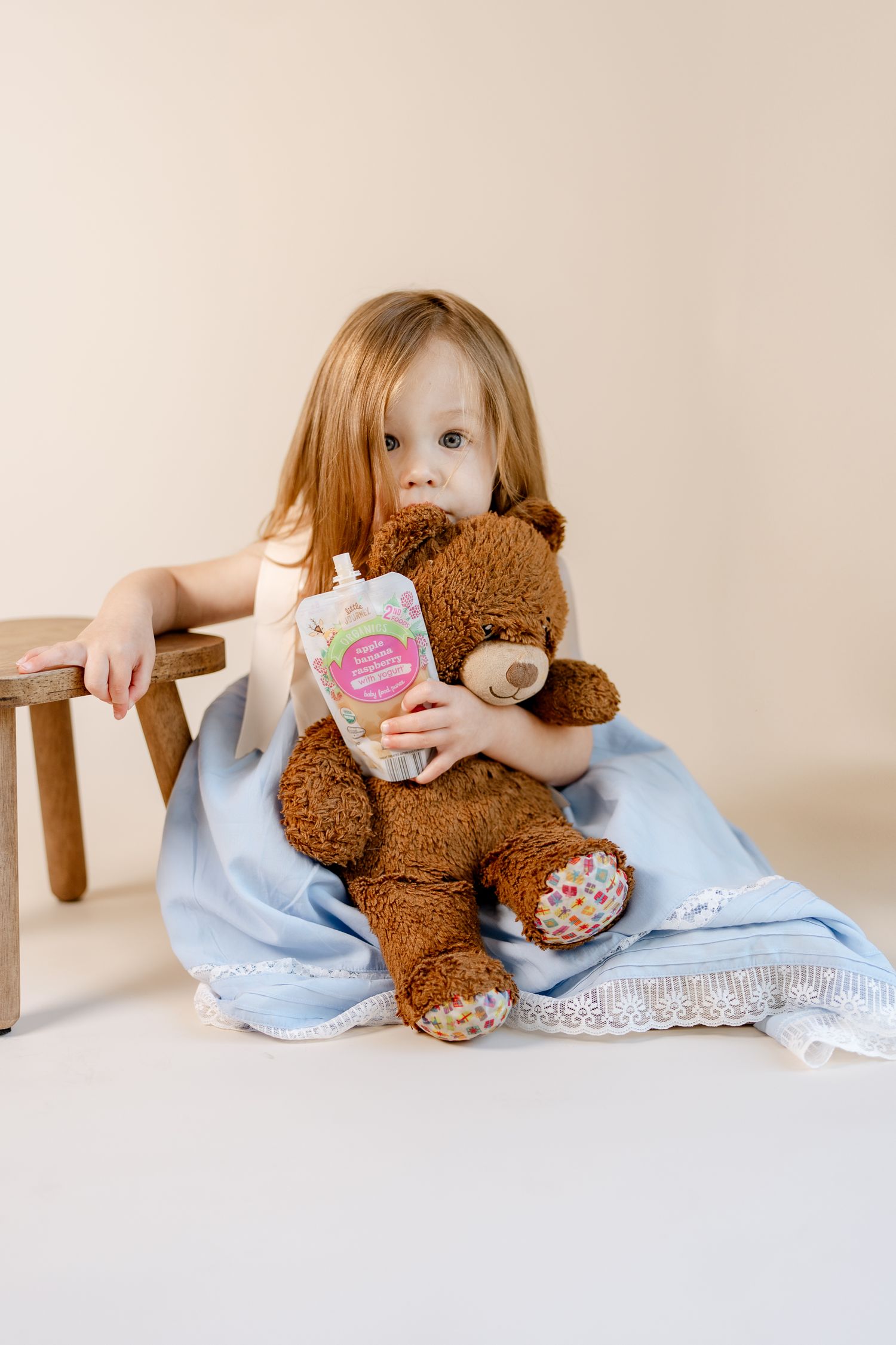A brown teddy bear rests beside a light blue lace-trimmed dress on a white surface.