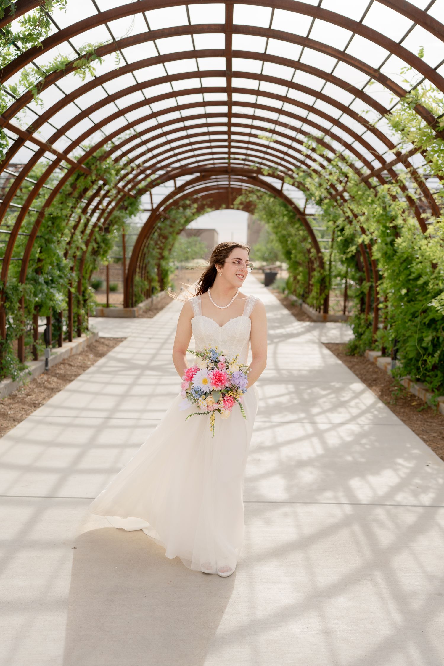 Bride in white gown holding colorful bouquet walks under arched garden trellis with lush greenery.