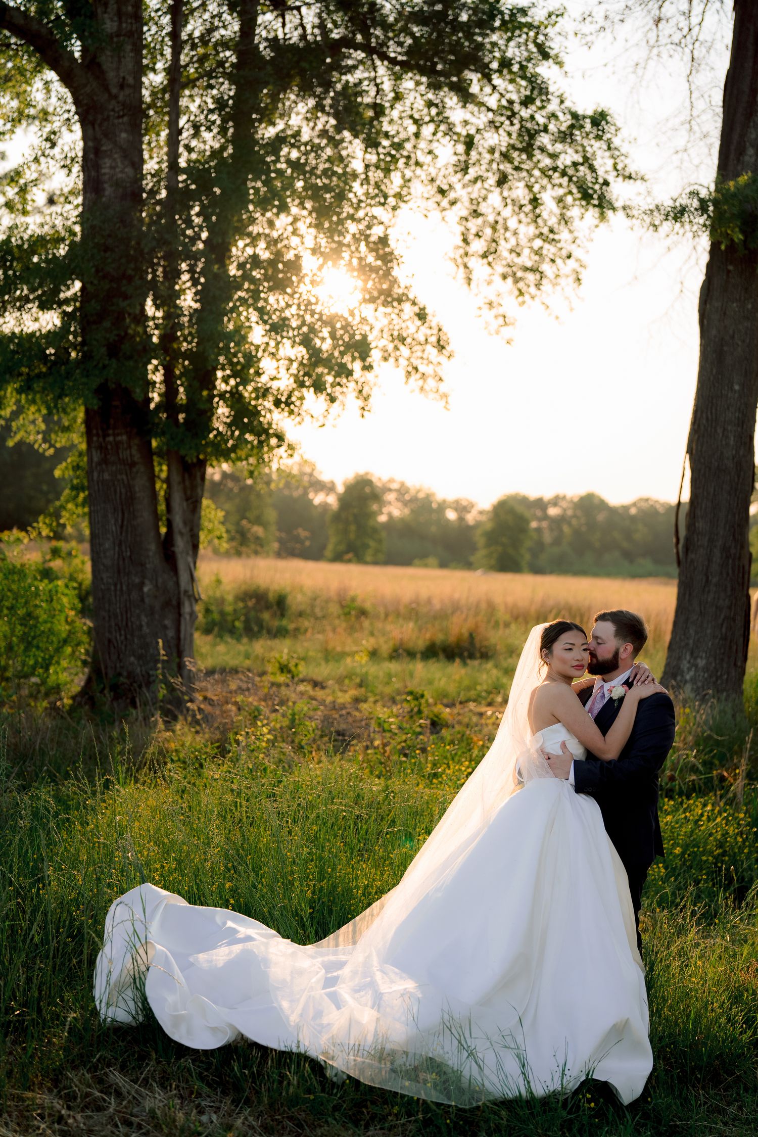 Bride and groom embracing at golden hour sunset in a lush green field with tall trees backdrop.