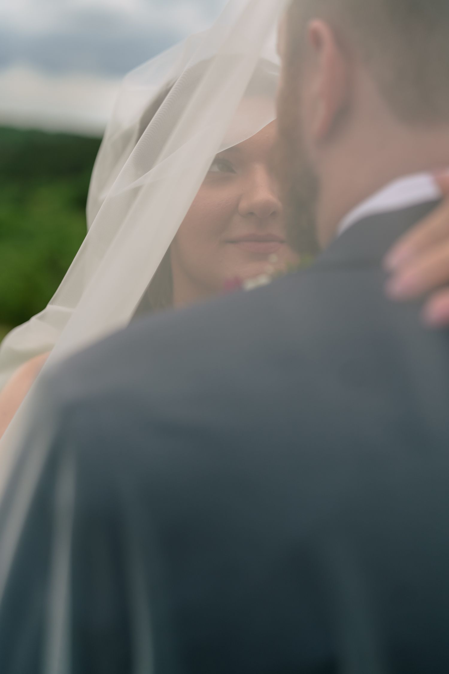 Bride smiling under flowing veil while embracing groom in gray suit during outdoor wedding portrait.