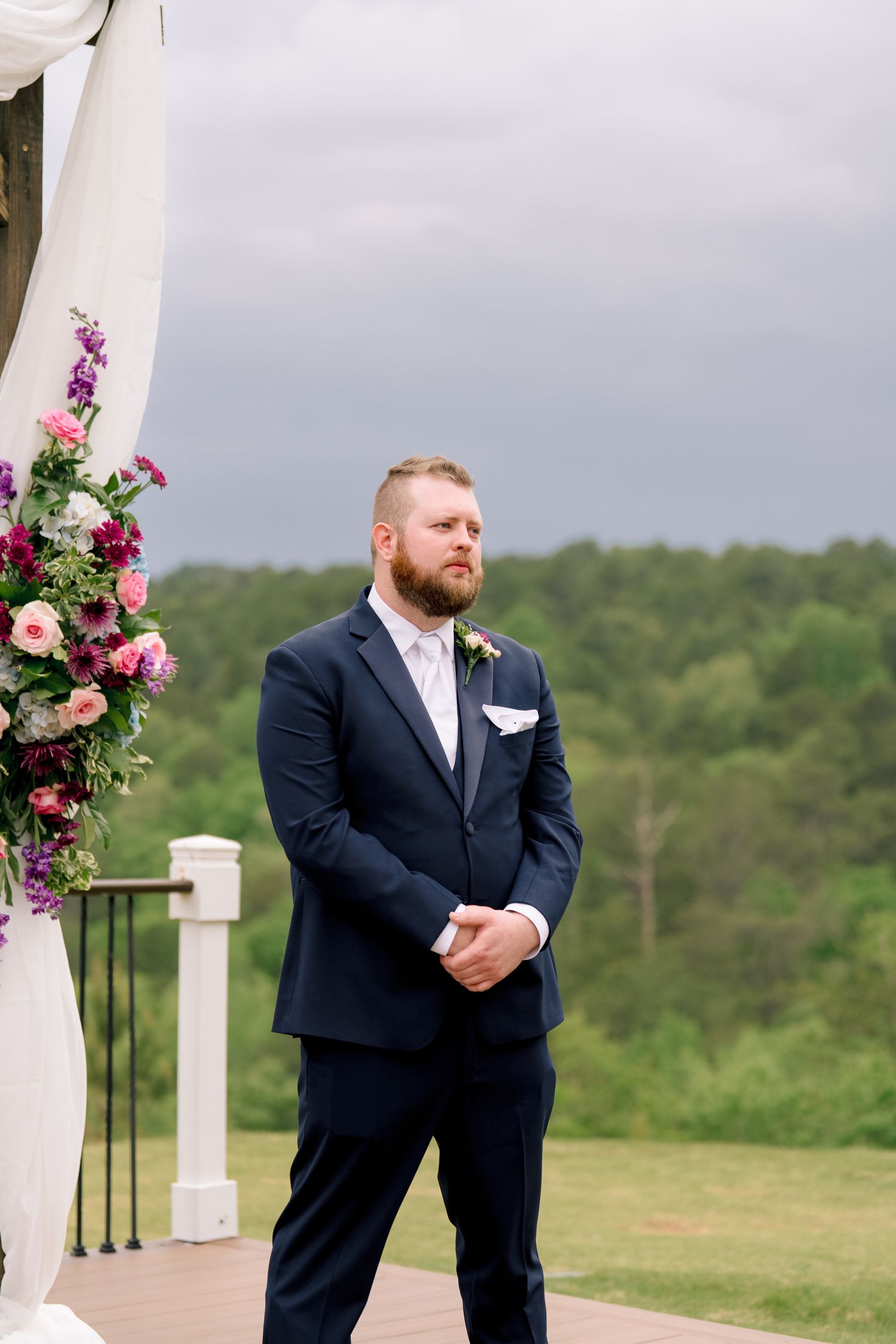 Groom in navy suit stands at outdoor wedding altar with floral arrangements under cloudy sky.