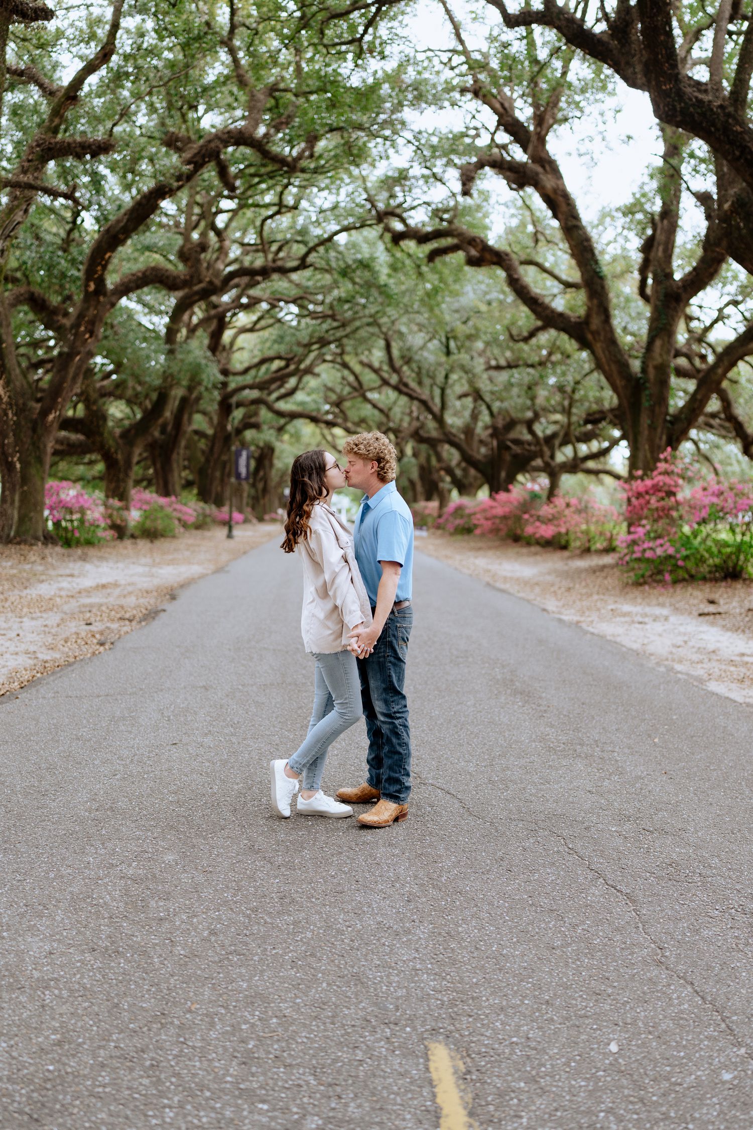 Couple stands together on path beneath canopy of oak trees and pink azaleas.