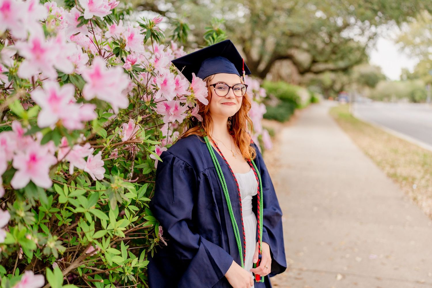 Graduate stands among pink azalea bushes wearing traditional academic regalia.