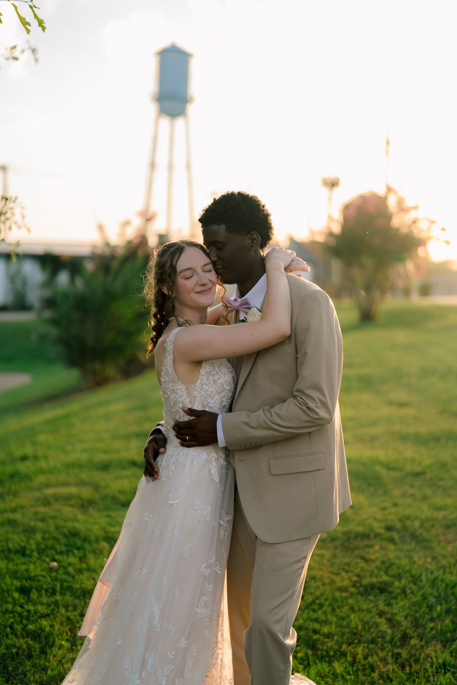 Interracial newlywed couple embracing at golden hour with a water tower in the background.