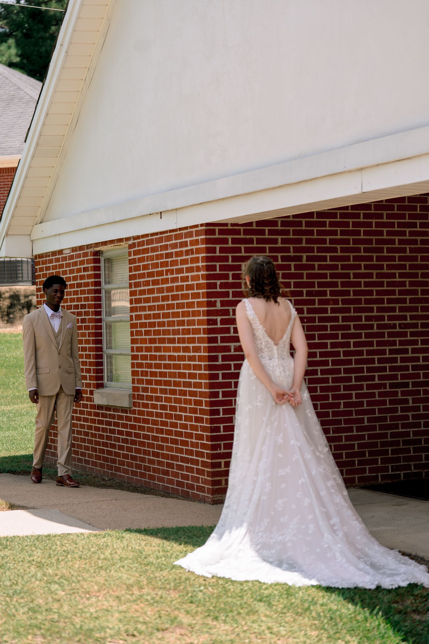 Bride in lace wedding gown stands outside brick church during first look moment with groom in tan suit.